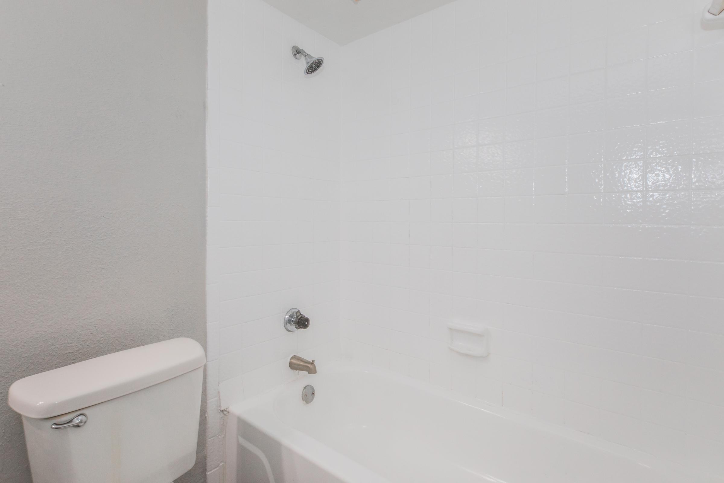 A clean, minimalist bathroom featuring a white tiled bathtub, a showerhead mounted on the wall, and a toilet to the left. The walls are plain and light-colored, enhancing the bright and uncluttered appearance of the space.