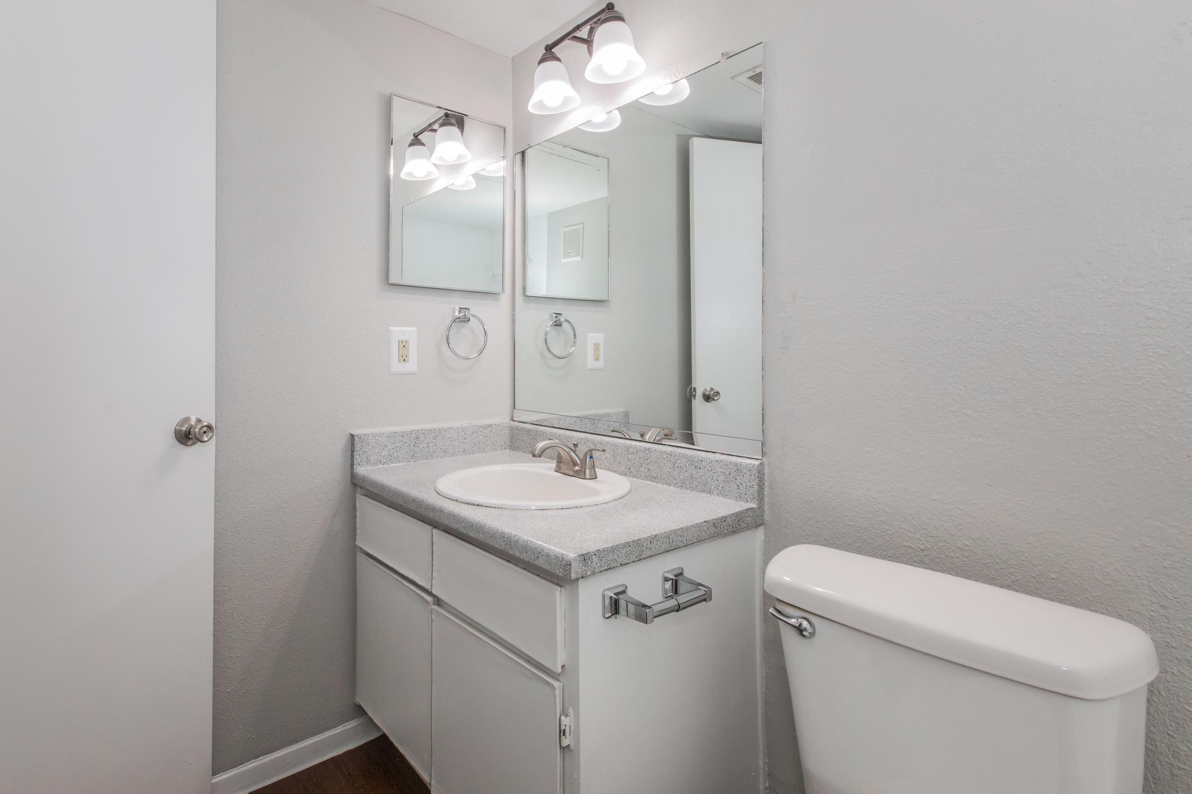 A modern bathroom featuring a white toilet, a double sink vanity with a gray countertop, and a large mirror above the sinks. The walls are painted in a light gray color, and there are three light fixtures above the mirror, providing bright illumination. The floor is dark wood, adding contrast to the overall decor.
