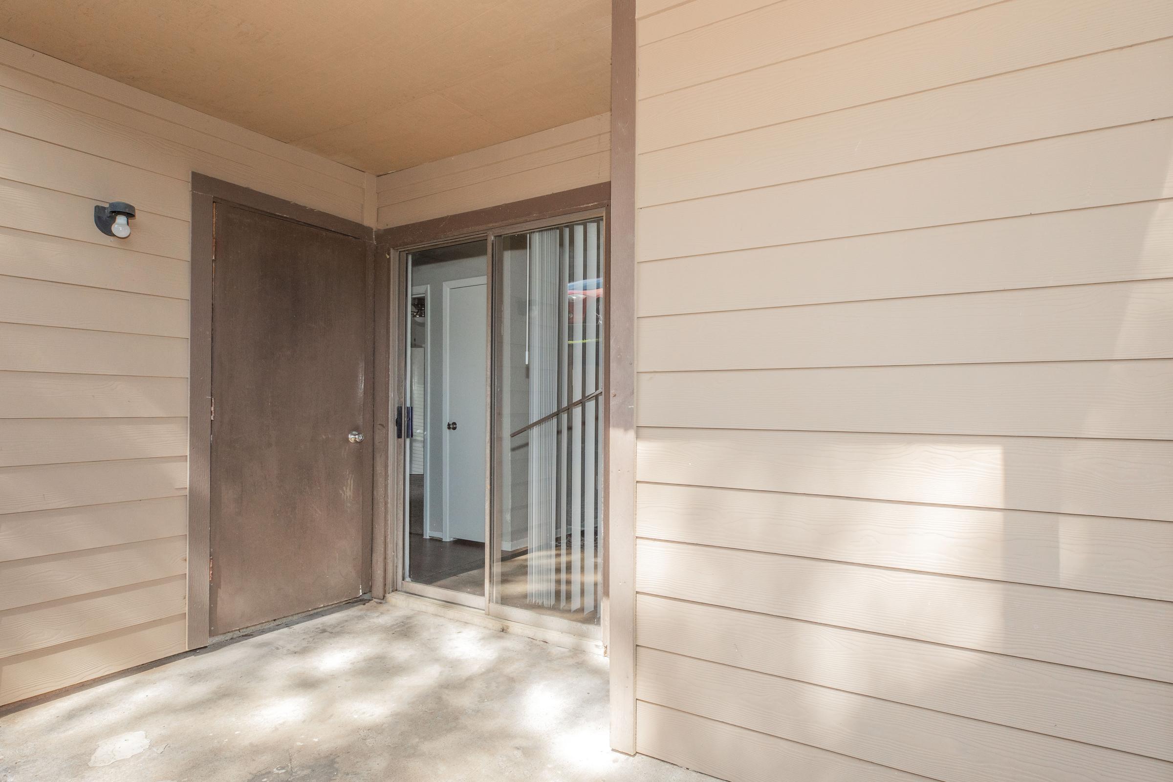 Entrance to a building featuring a sliding glass door and a solid wooden door. The walls are light beige, with a simple, clean aesthetic. The area has a small concrete space and is shaded, suggesting a welcoming entryway.