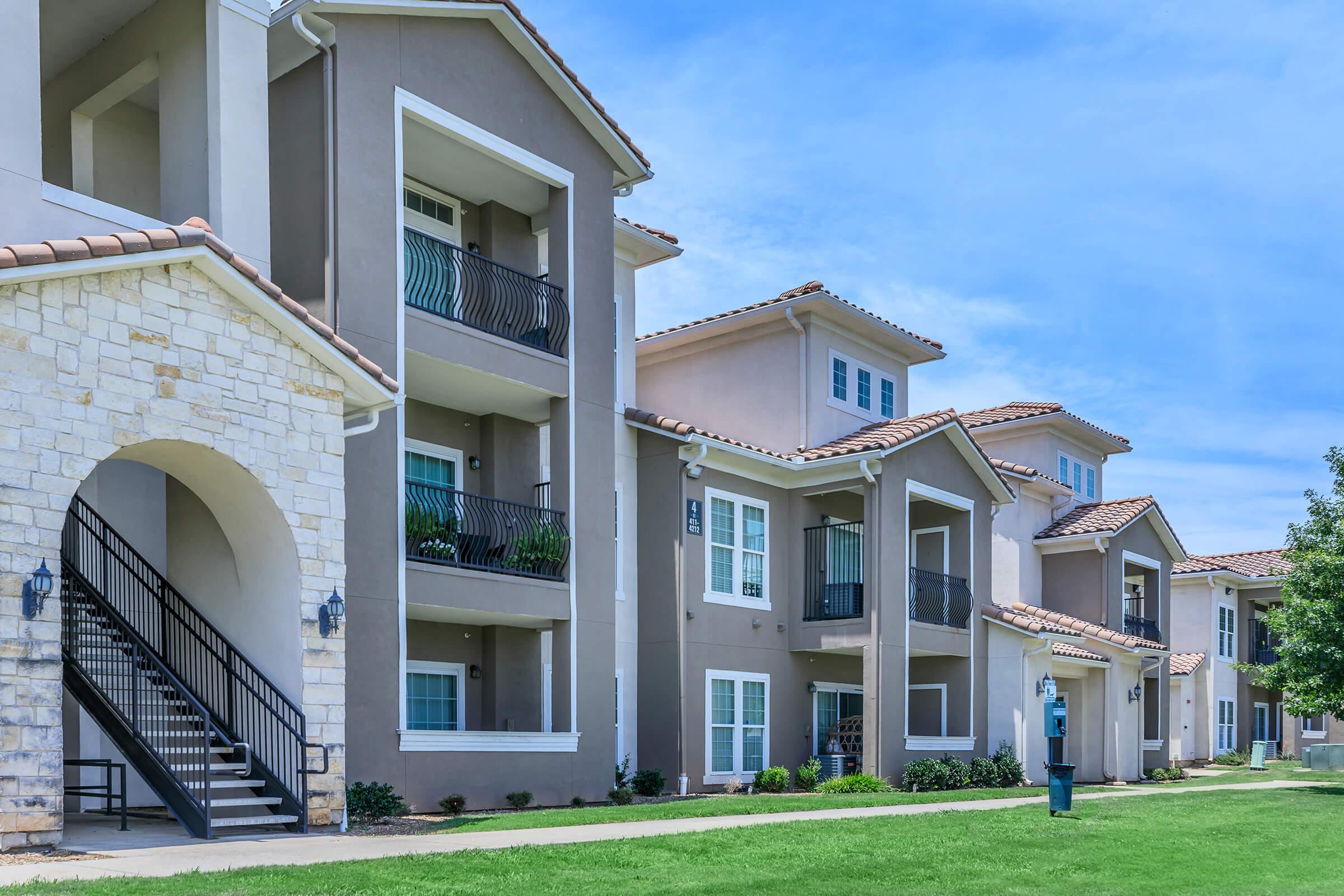 A row of modern residential apartments with stone and stucco facades under a clear blue sky. The buildings feature balconies, green landscaping, and a staircase leading to the upper levels. A well-maintained lawn is visible in the foreground, creating a welcoming atmosphere.