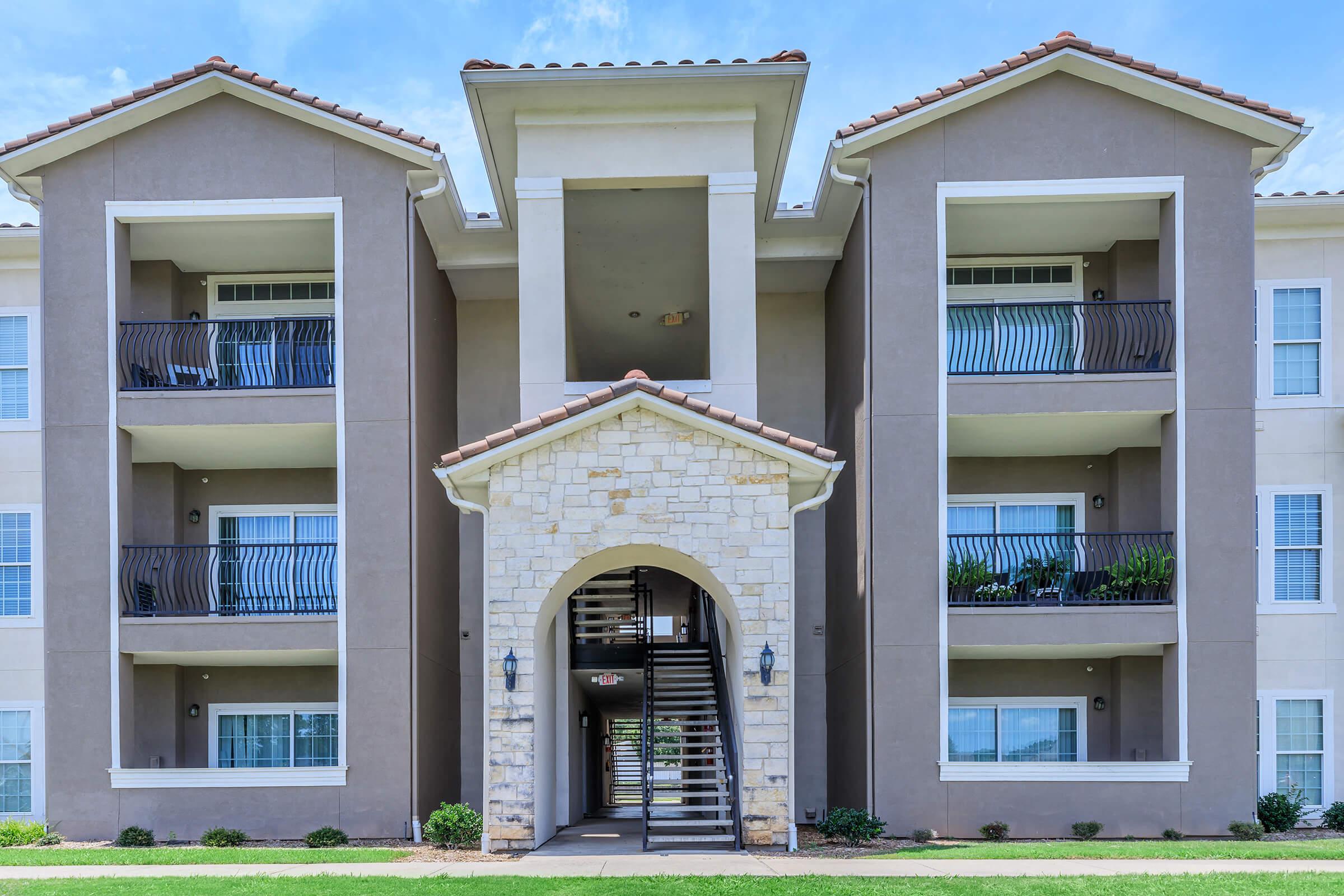 Front view of a multi-story apartment building featuring a central archway entrance, balconies on the upper floors, and a combination of stone and stucco exterior. A landscaped area is visible in the foreground, with a clear blue sky above.