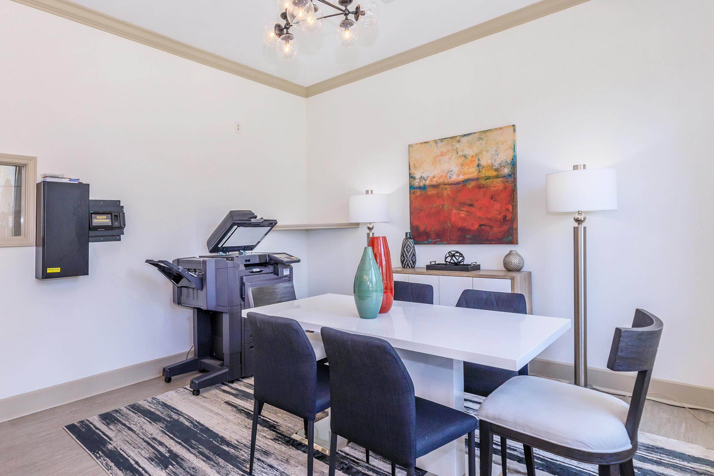 A modern dining area featuring a white table surrounded by black chairs, with a colorful abstract painting on the wall. There are two stylish lamps on either side of a light-colored sideboard, a decorative vase on the table, and a printer or copier in the corner. The flooring is light with a striped rug.