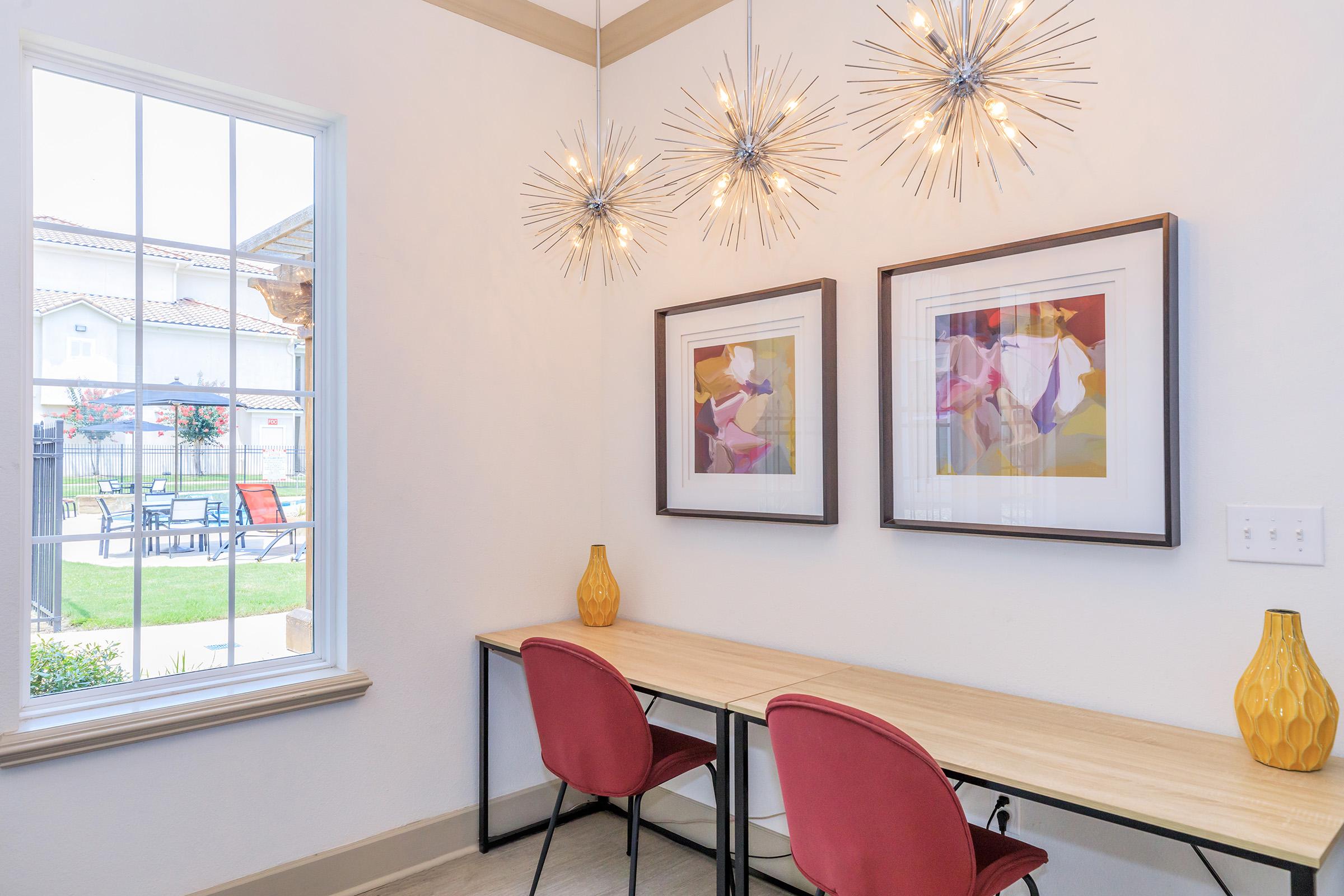 A well-lit modern home office features two red chairs at a wooden desk, with two framed abstract art pieces hanging on the wall. Sparkling starburst pendant lights hang from the ceiling, and a large window allows natural light to fill the space, offering a view of the outdoors and patio area.