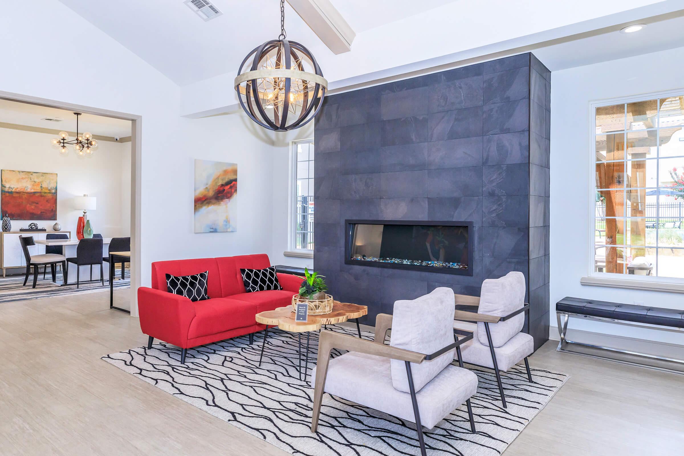 A modern living room featuring a red sofa, two light-colored chairs, and a stylish coffee table. The room includes a stone accent wall with a contemporary fireplace and a large pendant light fixture. Large windows allow natural light into the space, complemented by decorative artwork and a patterned area rug.