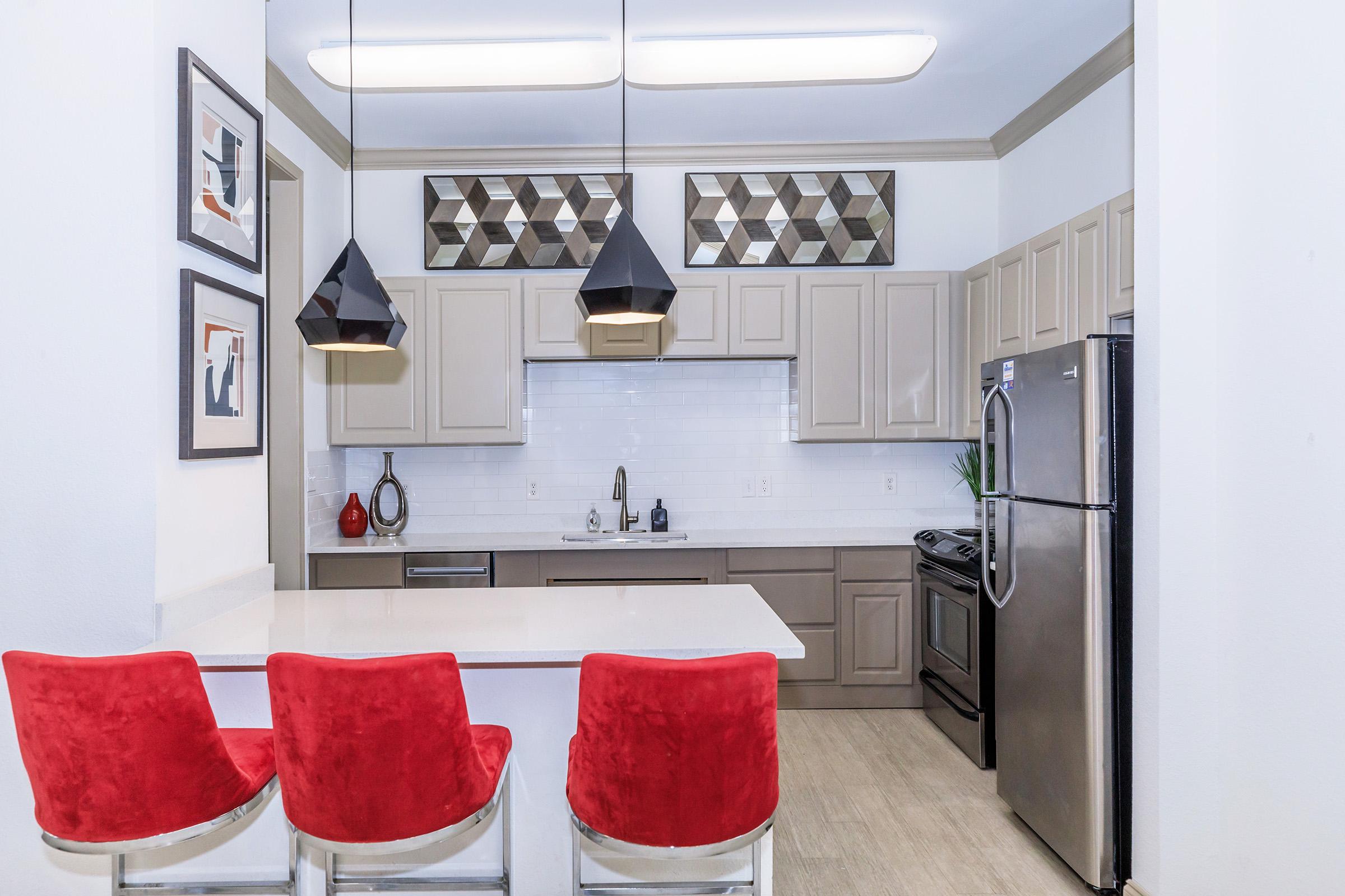 Modern kitchen featuring light-colored cabinetry, white tile backsplash, and stainless steel appliances. Three red upholstered bar stools are positioned at a counter, with decorative geometric artwork above the cabinets and stylish pendant lights hanging from the ceiling.