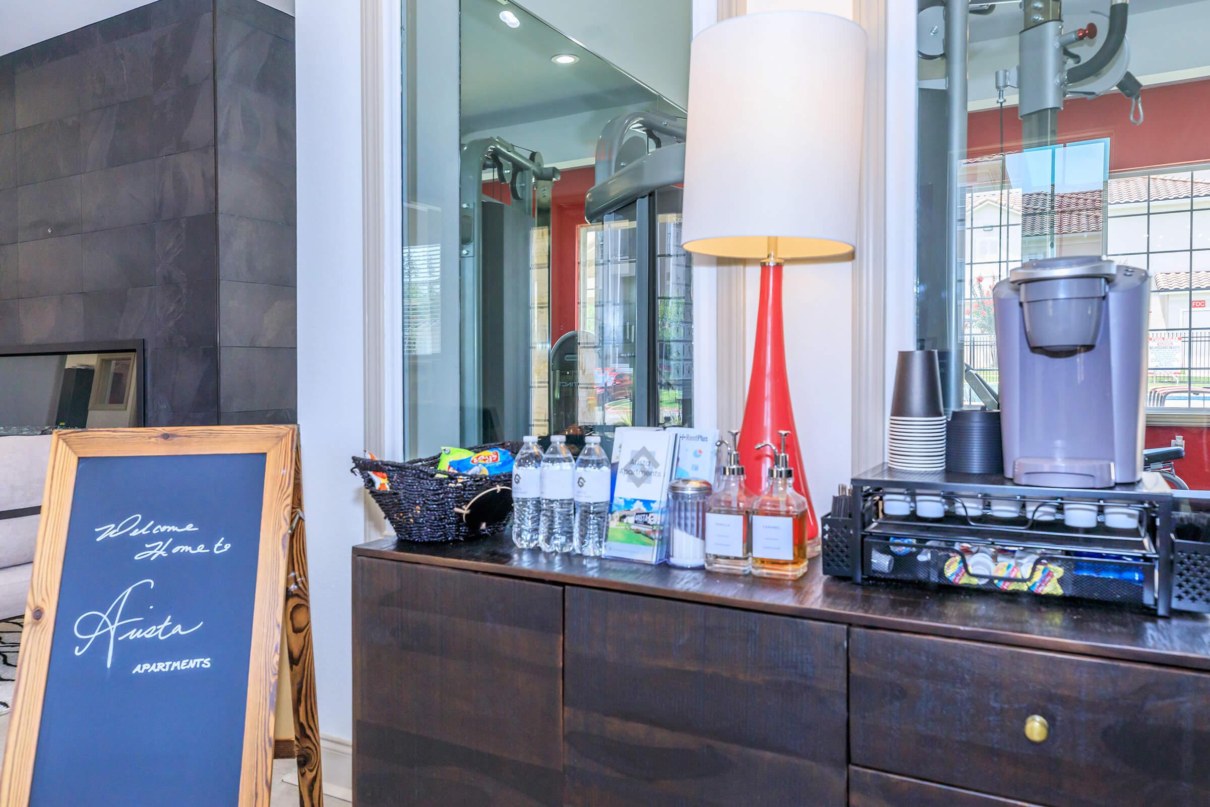 A modern reception area featuring a wooden chest with beverages including water bottles and coffee. A decorative lamp is positioned on the chest beside a sign welcoming guests. A chalkboard easel displays a handwritten message. The background shows a stylish interior with glass and dark walls.