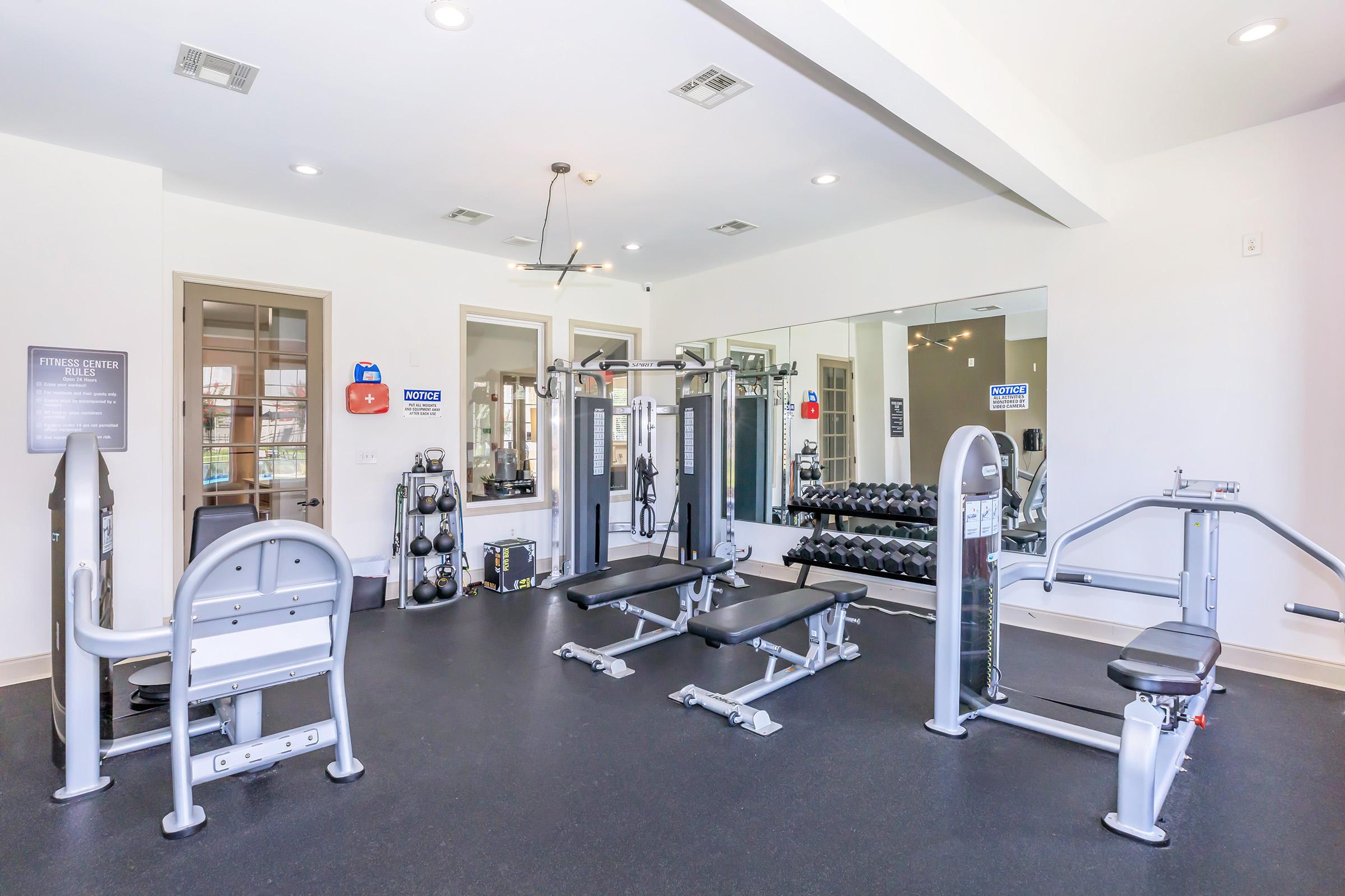 A well-lit gym area featuring various exercise equipment, including weight machines, free weights, and benches. Large mirrors are mounted on the wall, and there are fitness posters. The space is clean and organized, promoting an inviting atmosphere for workouts.