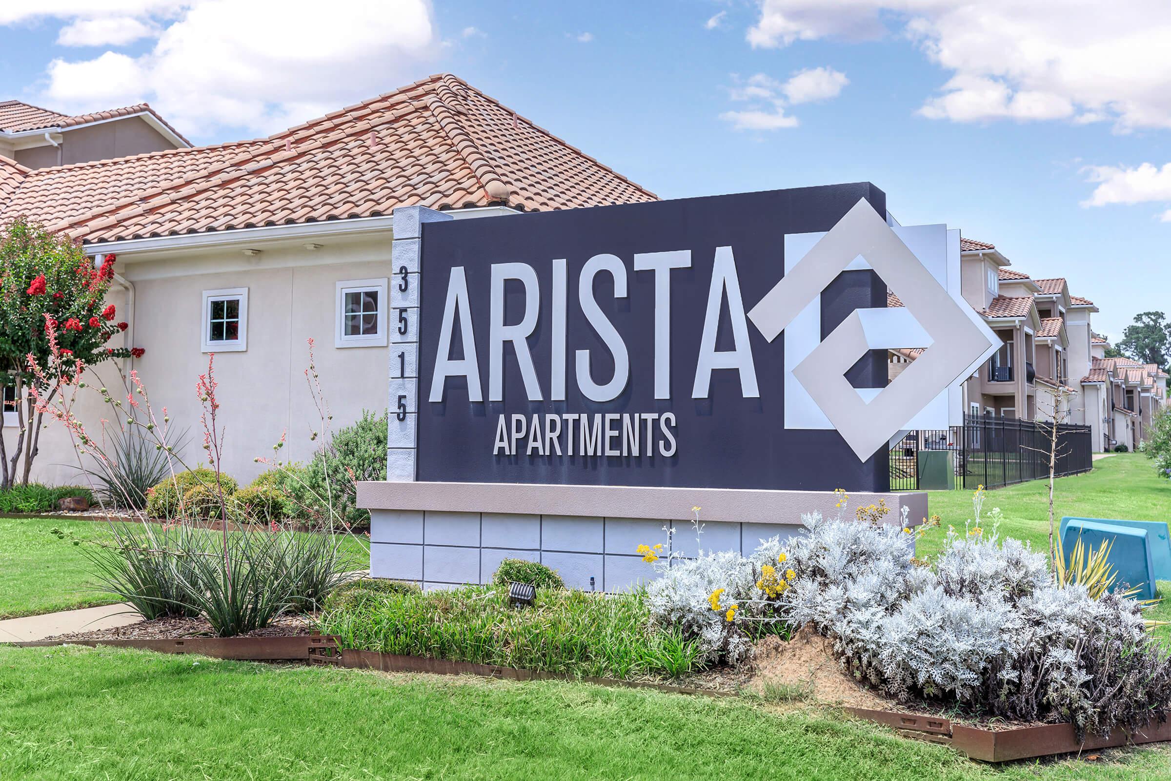 Sign for Arista Apartments displaying the name prominently. The sign is located in a landscaped area with grass and various plants. In the background, there are residential buildings with tiled roofs under a partly cloudy sky.