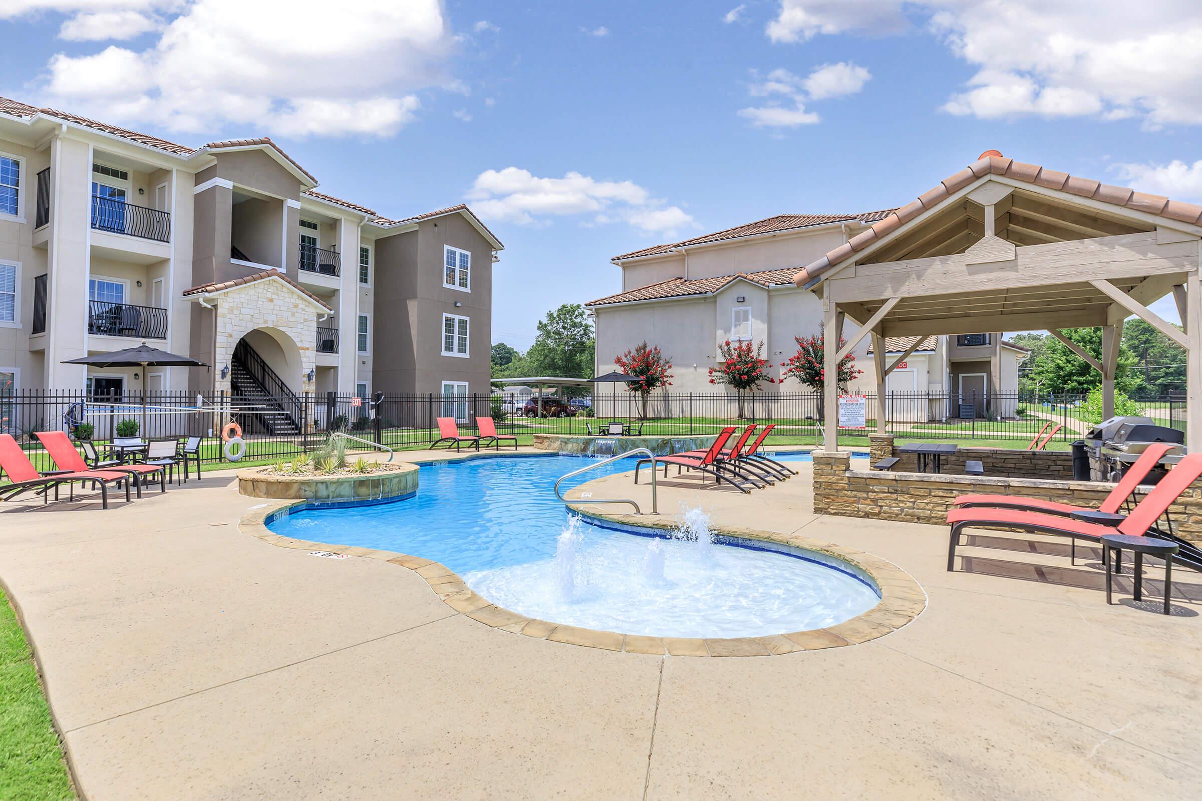 Swimming pool with a curved design surrounded by lounge chairs and a gazebo. In the background, there are two residential buildings with balconies. The area is lushly landscaped with trees and colorful flowers under a clear blue sky, creating a relaxing outdoor environment.