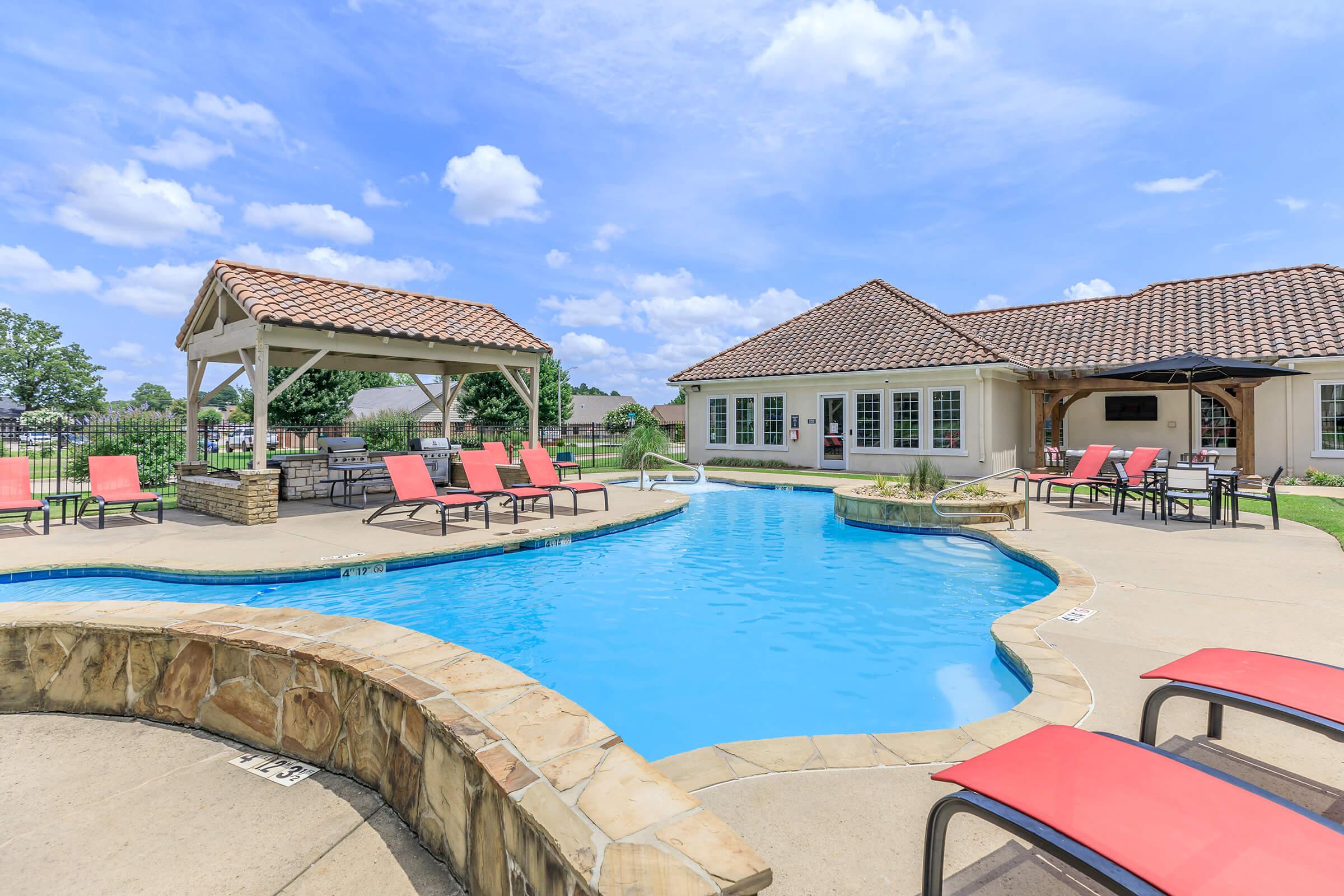 A swimming pool surrounded by lounge chairs and a shaded gazebo. The pool features a curved edge and is complemented by a stone deck. In the background, there's a building with large windows and a tile roof, under a bright blue sky with fluffy clouds.