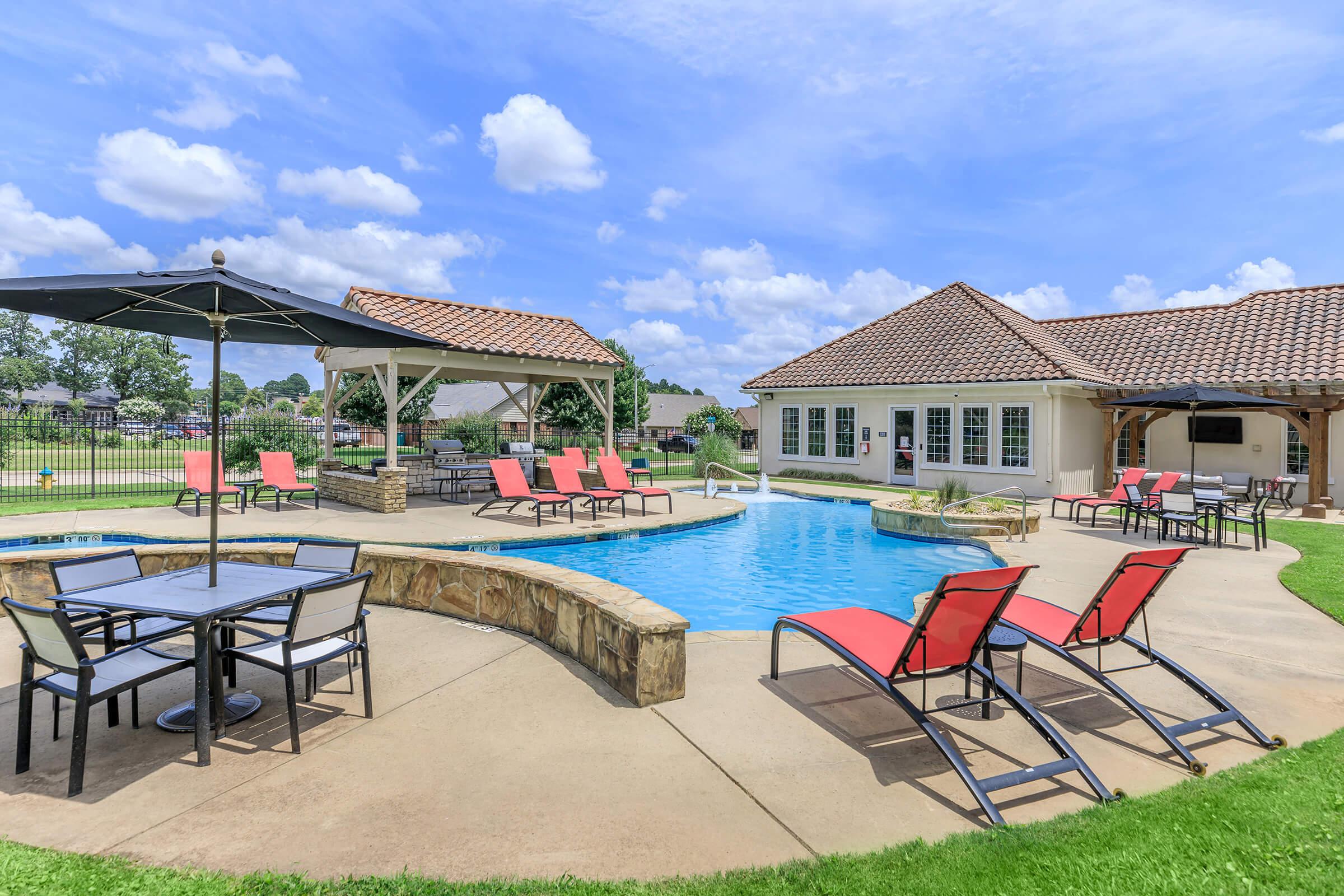A bright outdoor pool area featuring a rectangular swimming pool with lounge chairs and tables around it. There is a shaded patio with umbrellas, surrounded by green grass and trees in the background, under a partly cloudy blue sky. The area is ideal for relaxation and leisure activities.