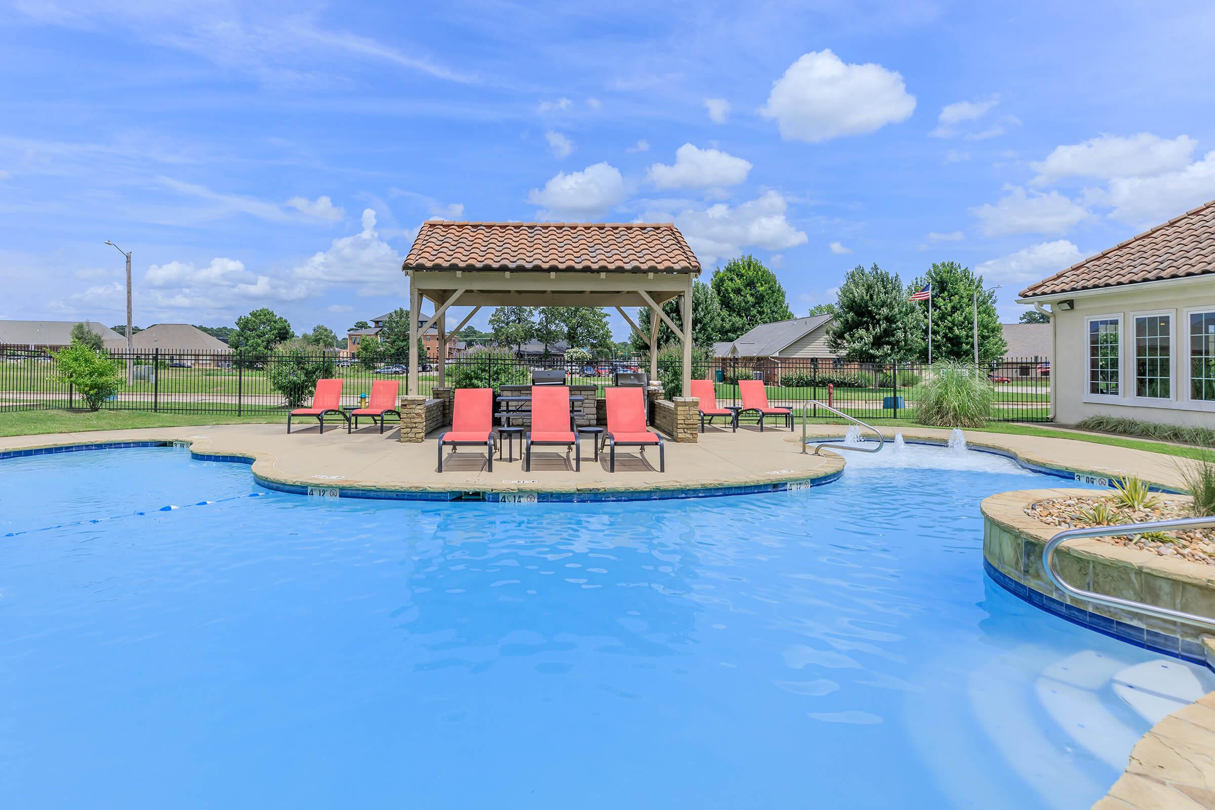 A spacious pool area featuring a clear blue swimming pool surrounded by lounge chairs. In the center, there's a shaded gazebo. Lush green trees and homes can be seen in the background under a bright blue sky with fluffy white clouds.