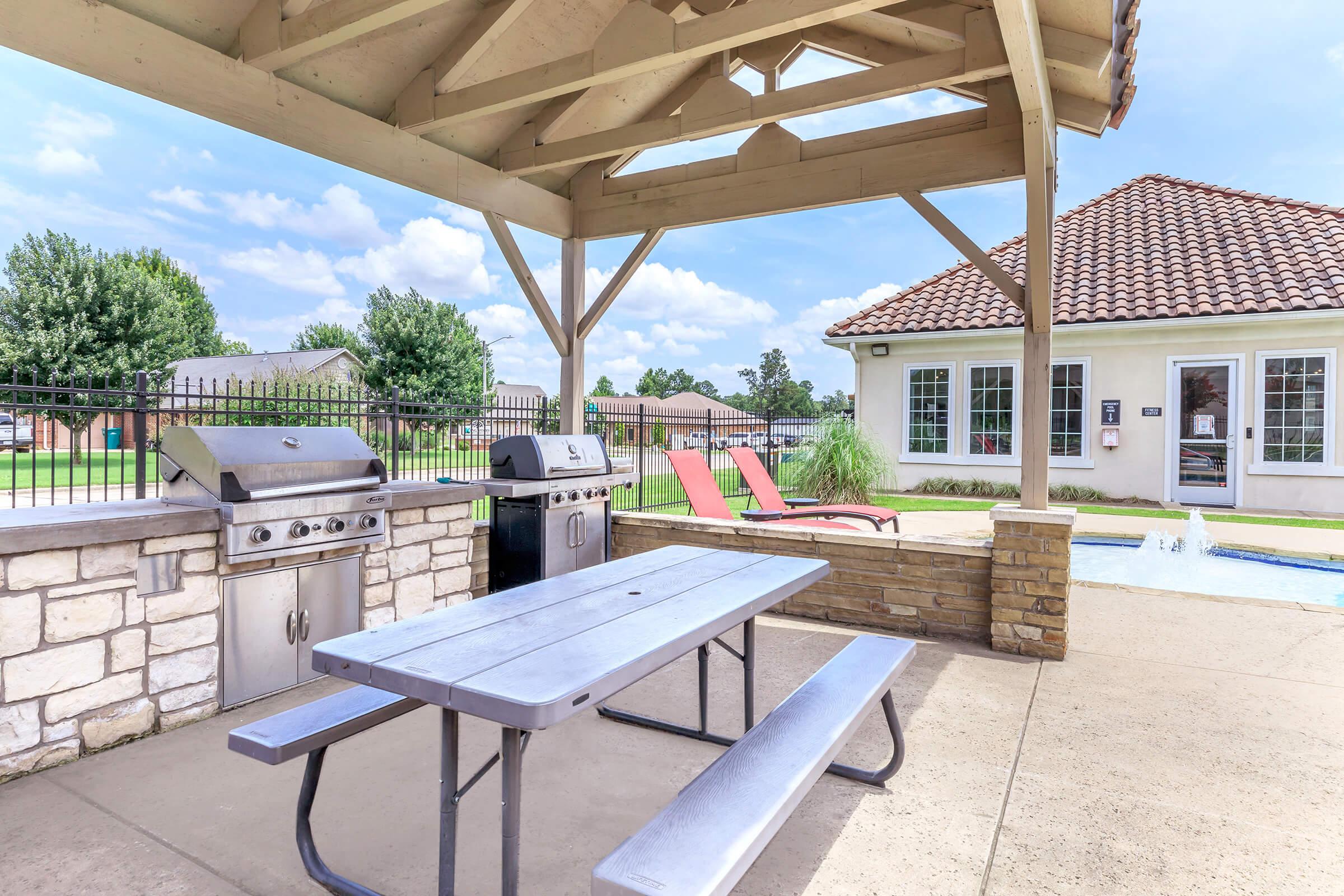 Outdoor barbecue area featuring a picnic table, two grilling stations, and red lounge chairs under a wooden pavilion. In the background, there is a swimming pool and a residential building, surrounded by green grass and trees, with a clear blue sky above.