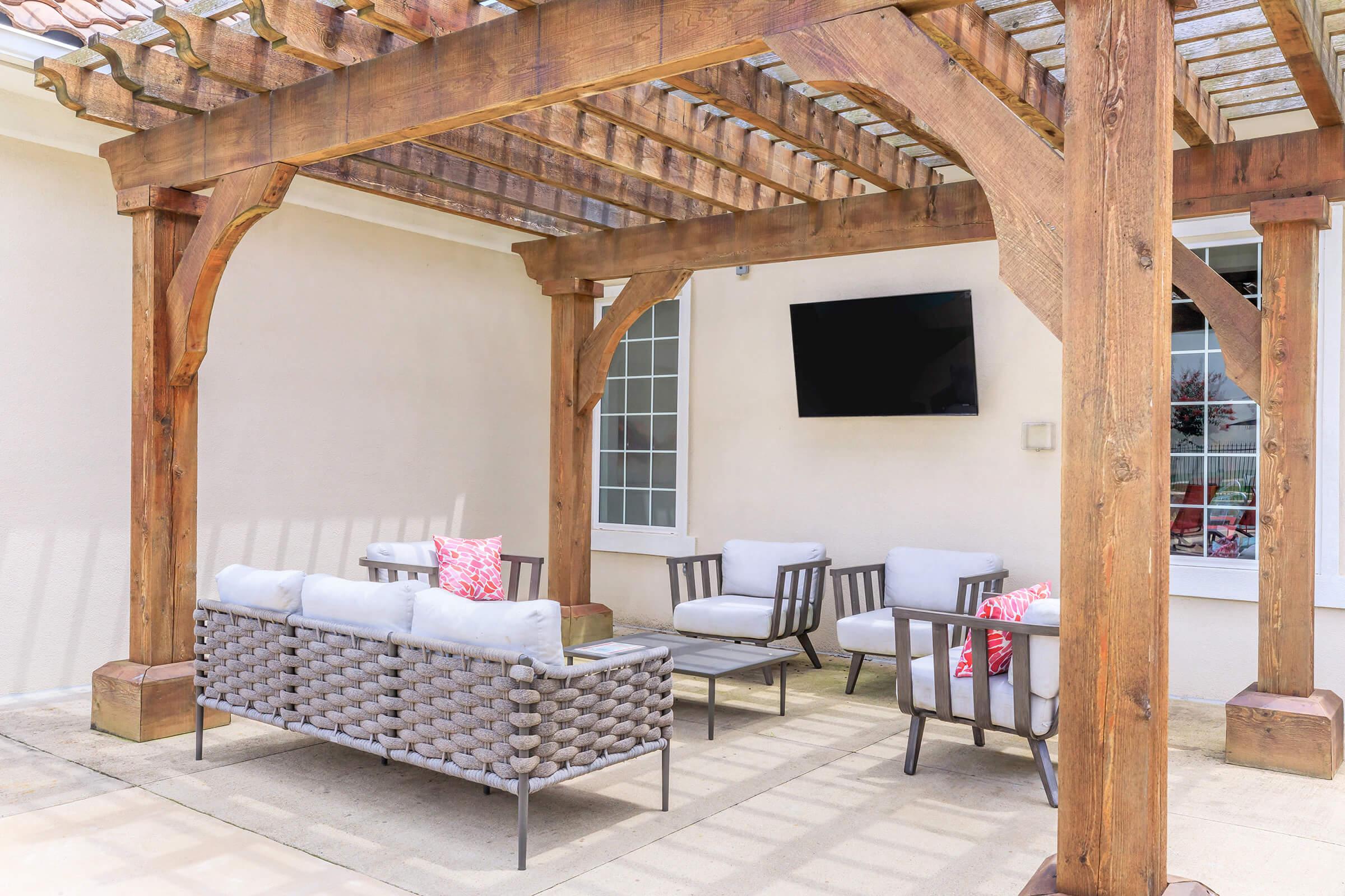 A spacious outdoor seating area featuring a wooden pergola, comfortable wicker furniture with cushions, and a mounted TV on the wall. The setting is well-lit, with natural light illuminating the space, creating a cozy atmosphere for relaxation or entertainment.