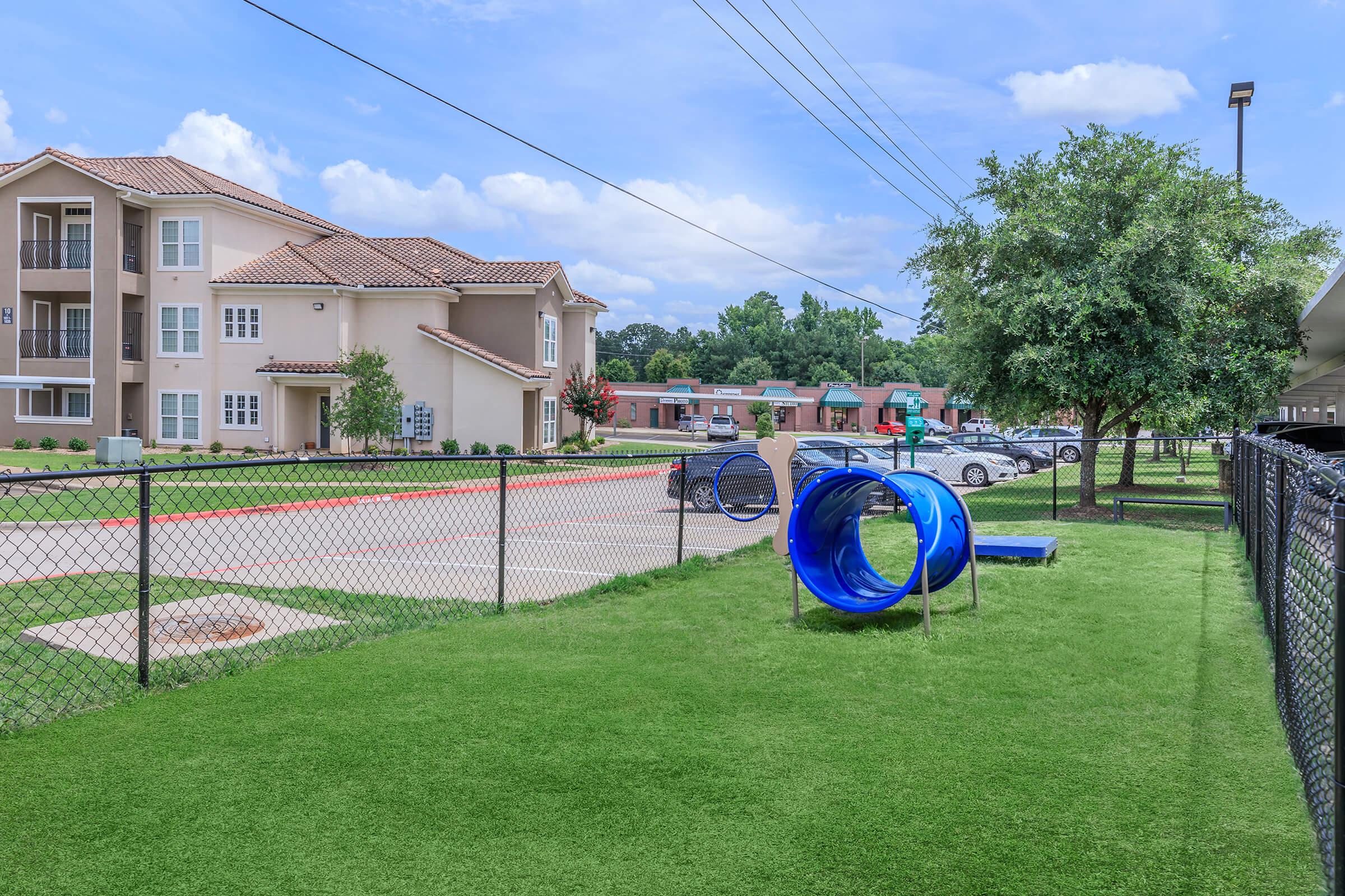 View of a residential area featuring a playground with a blue slide on green grass. In the background, there are apartment buildings and cars parked along a road. The sky is partly cloudy, adding a bright atmosphere to the scene. A fence surrounds the playground area.
