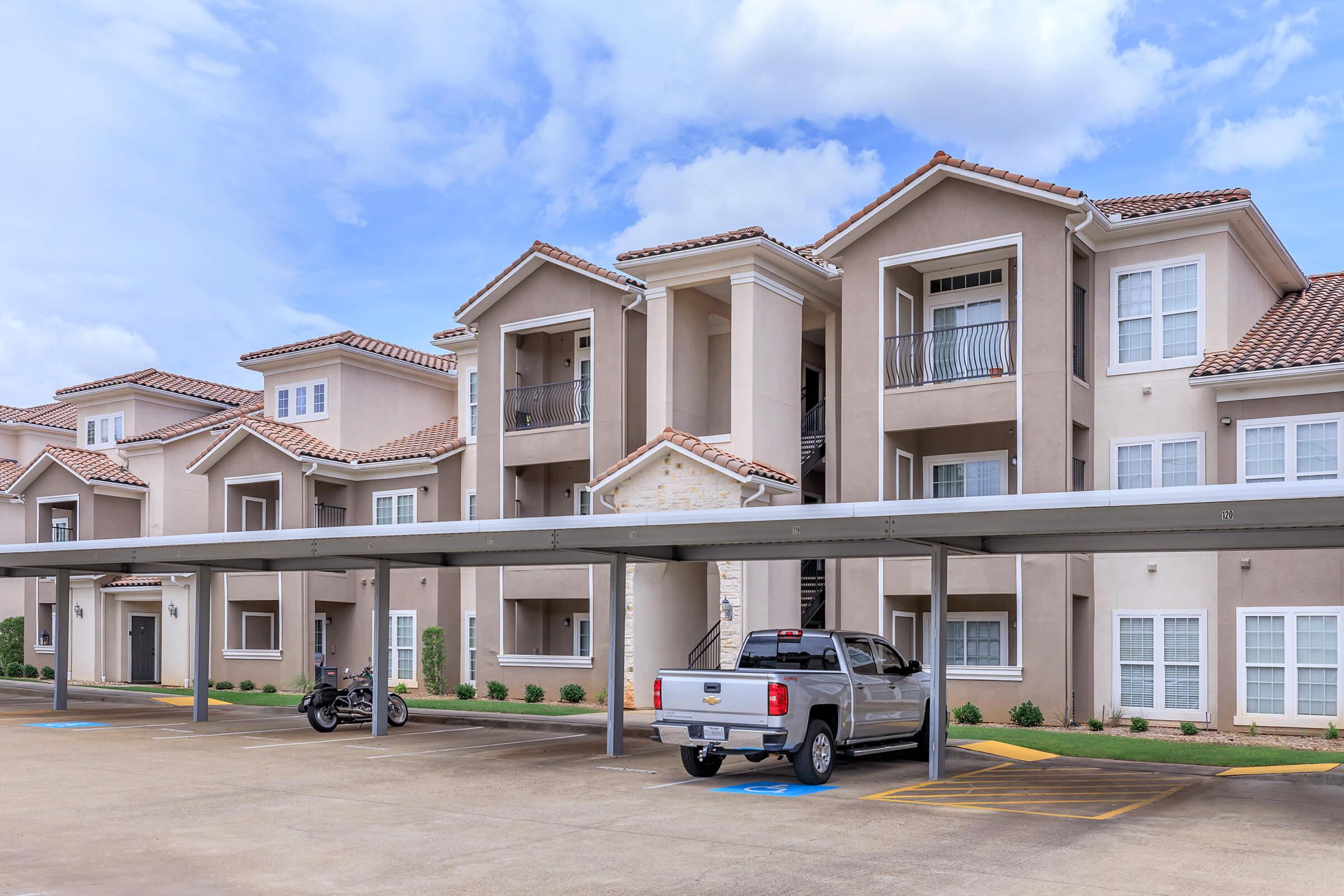 A modern apartment complex with several three-story buildings featuring light-colored exterior walls and tile roofs. There is a covered parking area in the foreground, with a white pickup truck and a motorcycle parked underneath. The sky is partly cloudy, adding a bright atmosphere to the scene.
