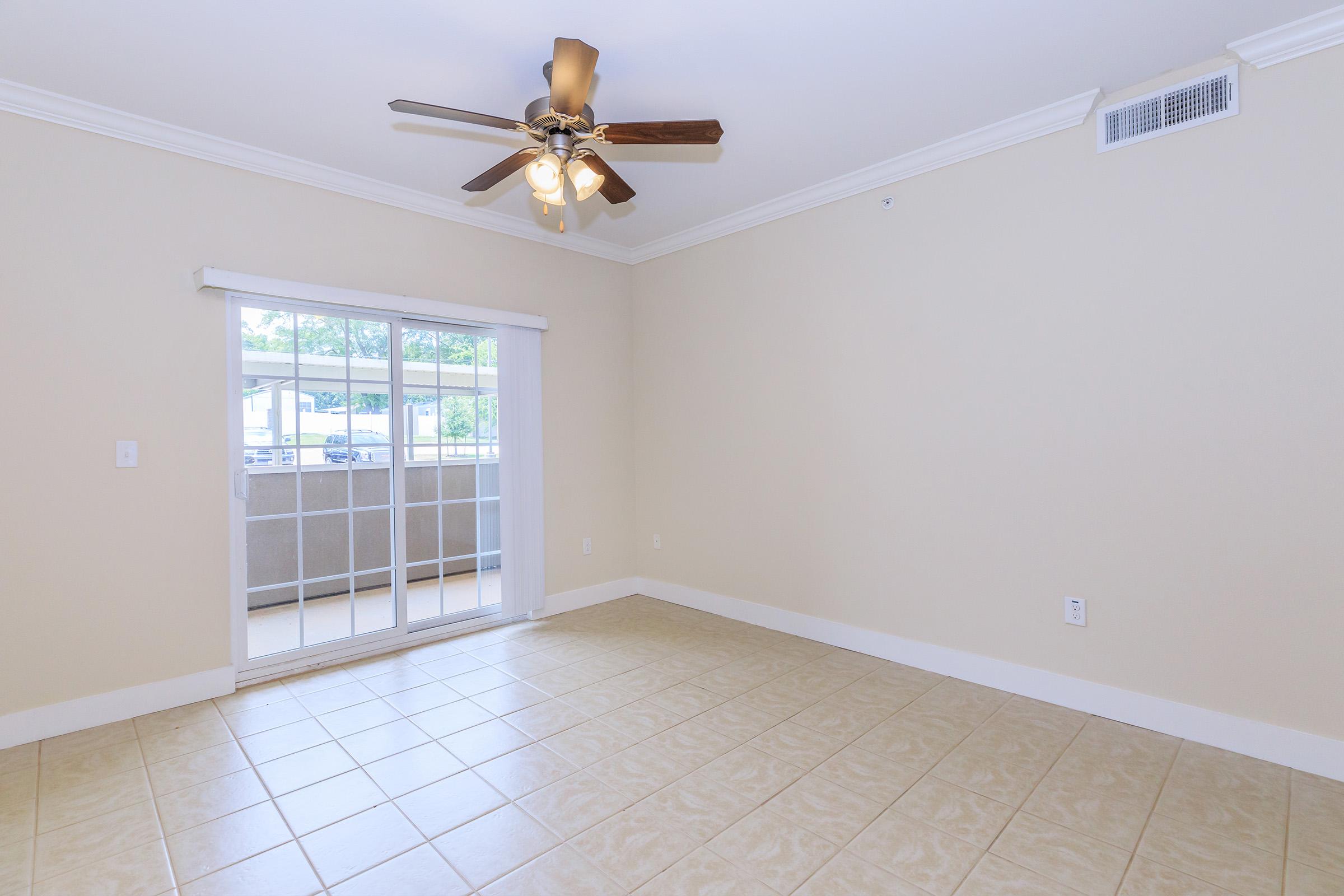 Empty room with light beige walls and tiled flooring. A ceiling fan with three bulbs hangs from the ceiling. A sliding glass door leads to a balcony, letting in natural light. The room has a simple, spacious layout, with no furniture present.