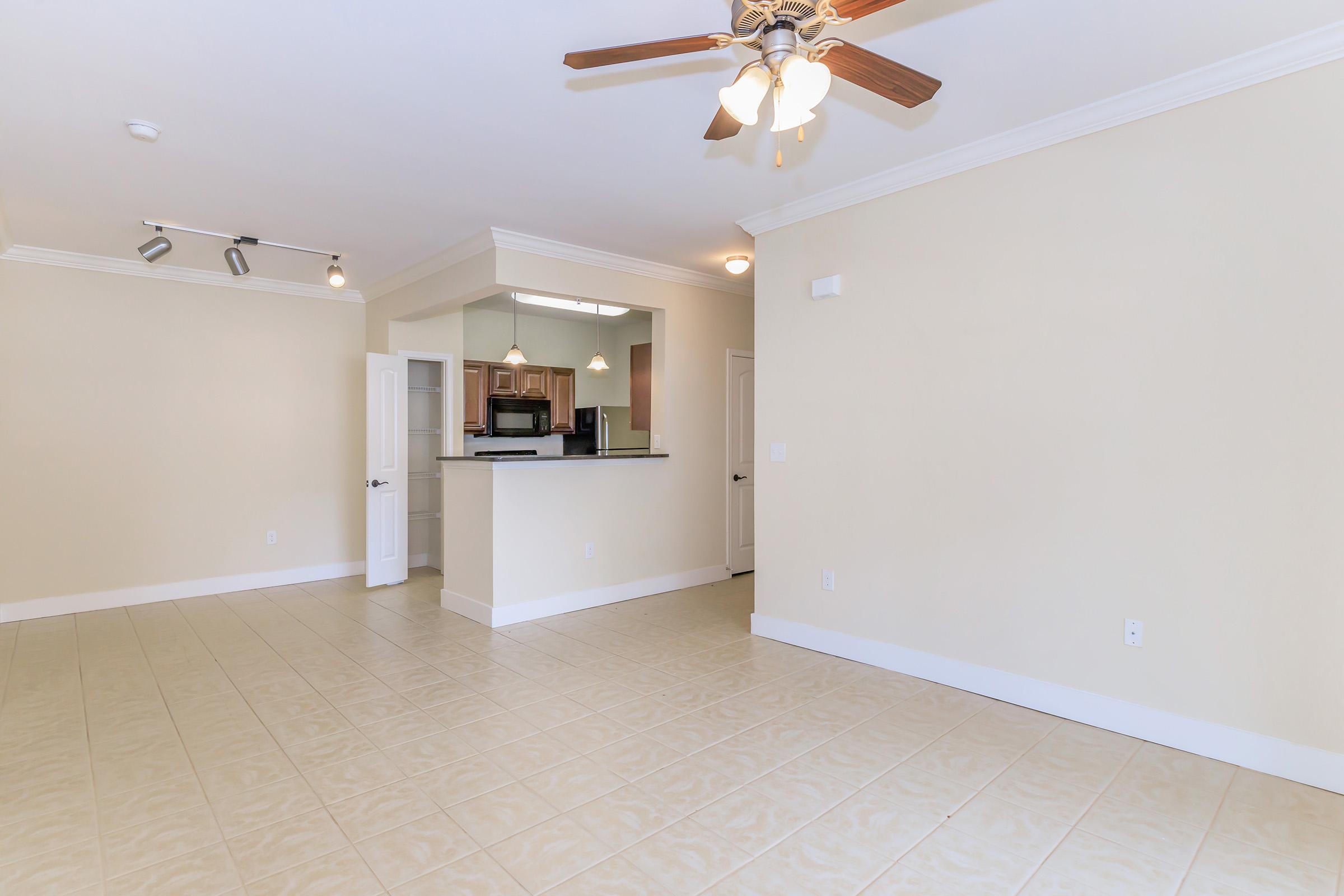 A spacious, empty room featuring light-colored walls and tiled flooring. There is a ceiling fan with lights, and a doorway leads to a kitchen area with dark cabinetry visible. The space is well-lit, with natural light coming from the doorway, creating an inviting atmosphere.