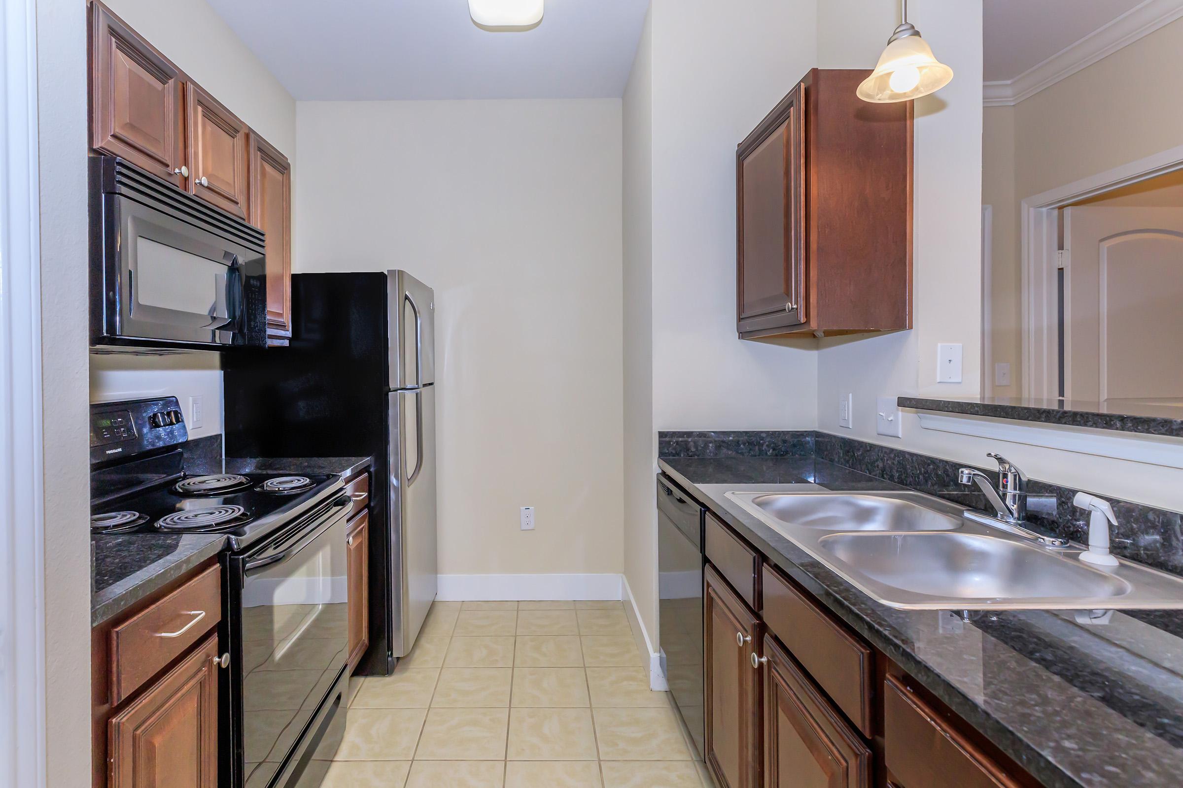 A modern kitchen featuring dark wood cabinetry, black appliances including a stove, microwave, and refrigerator, and a double sink. The countertops are dark granite, and the floor is tiled. The space is well-lit with overhead lighting and has a simple, clean design.