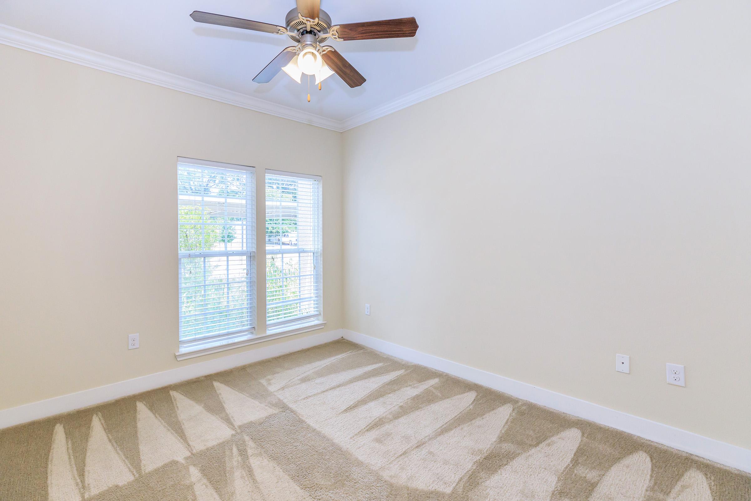 Bright and spacious empty room with a ceiling fan, featuring large windows that let in natural light. The floor is covered with beige carpet, and the walls are painted a soft cream color. Shadows from the fan blades are cast on the carpet, adding depth to the serene atmosphere.