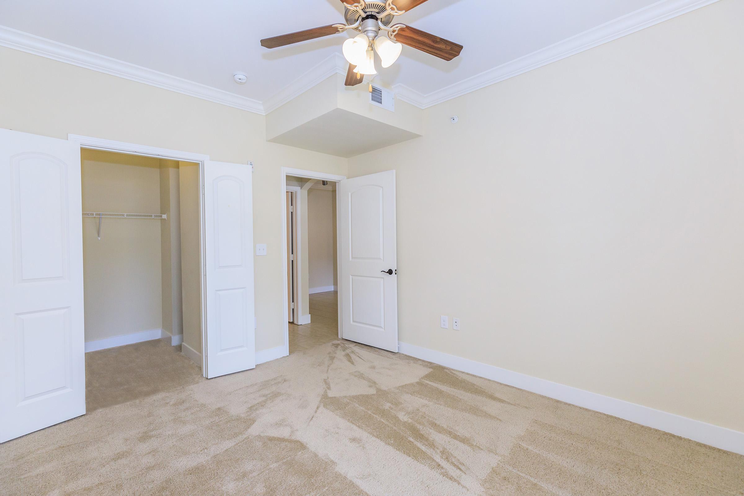A spacious room featuring light beige walls and carpet, with a ceiling fan. There are two white doors: one leading to a small closet and the other potentially to another room or hallway. The room is well-lit and appears empty, creating an inviting and fresh atmosphere.