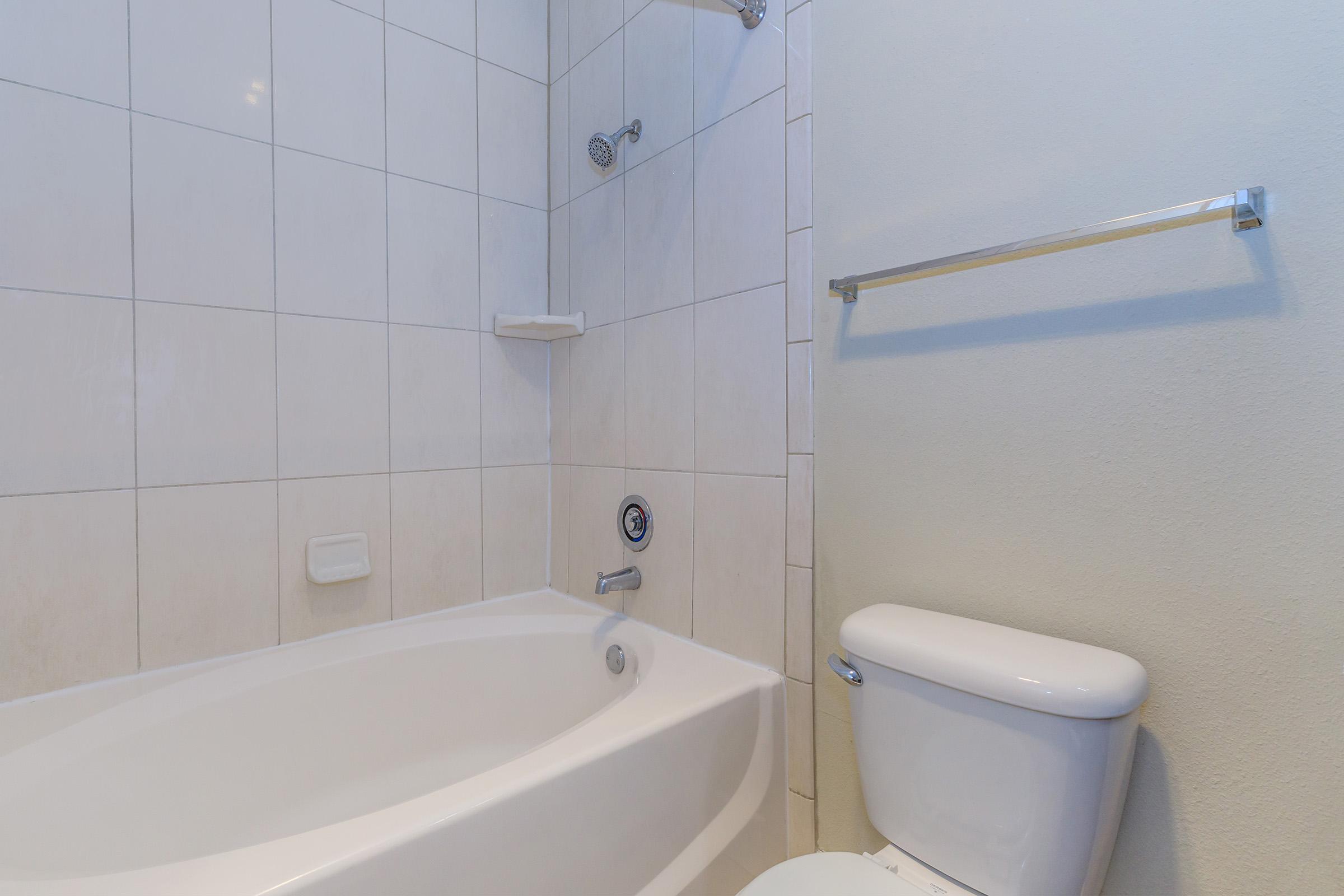 A clean bathroom featuring a white bathtub with a showerhead above, white tiled walls, a small shelf, and a toilet nearby. The overall design is modern and minimalist, creating a functional space for bathing and hygiene.