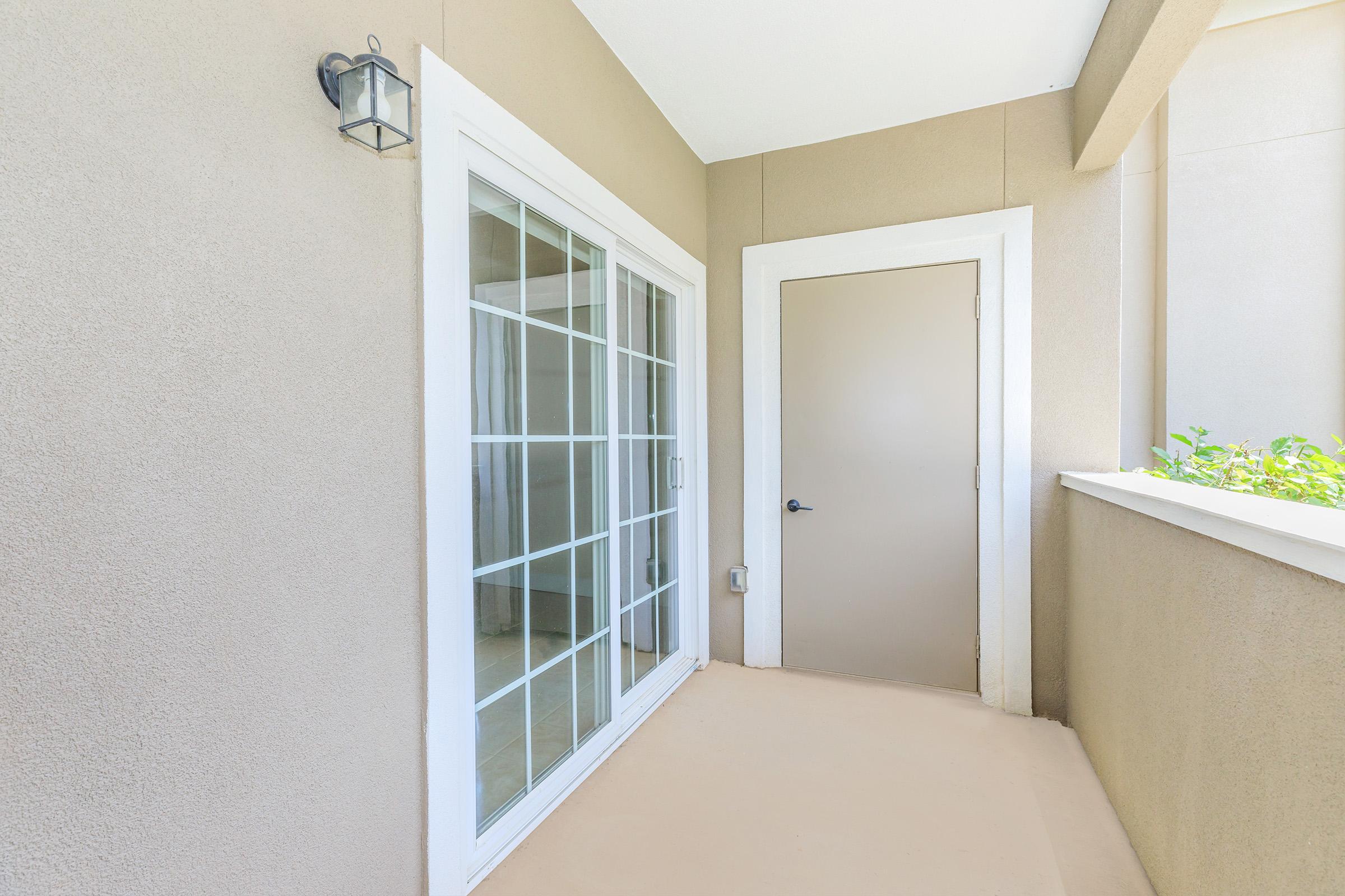 A well-lit exterior view of a small balcony area featuring a glass sliding door and a plain wall with a light fixture. There is a solid door to the right, and the corner has some greenery visible, providing a fresh touch to the space. The floor is neutral-colored and clean.