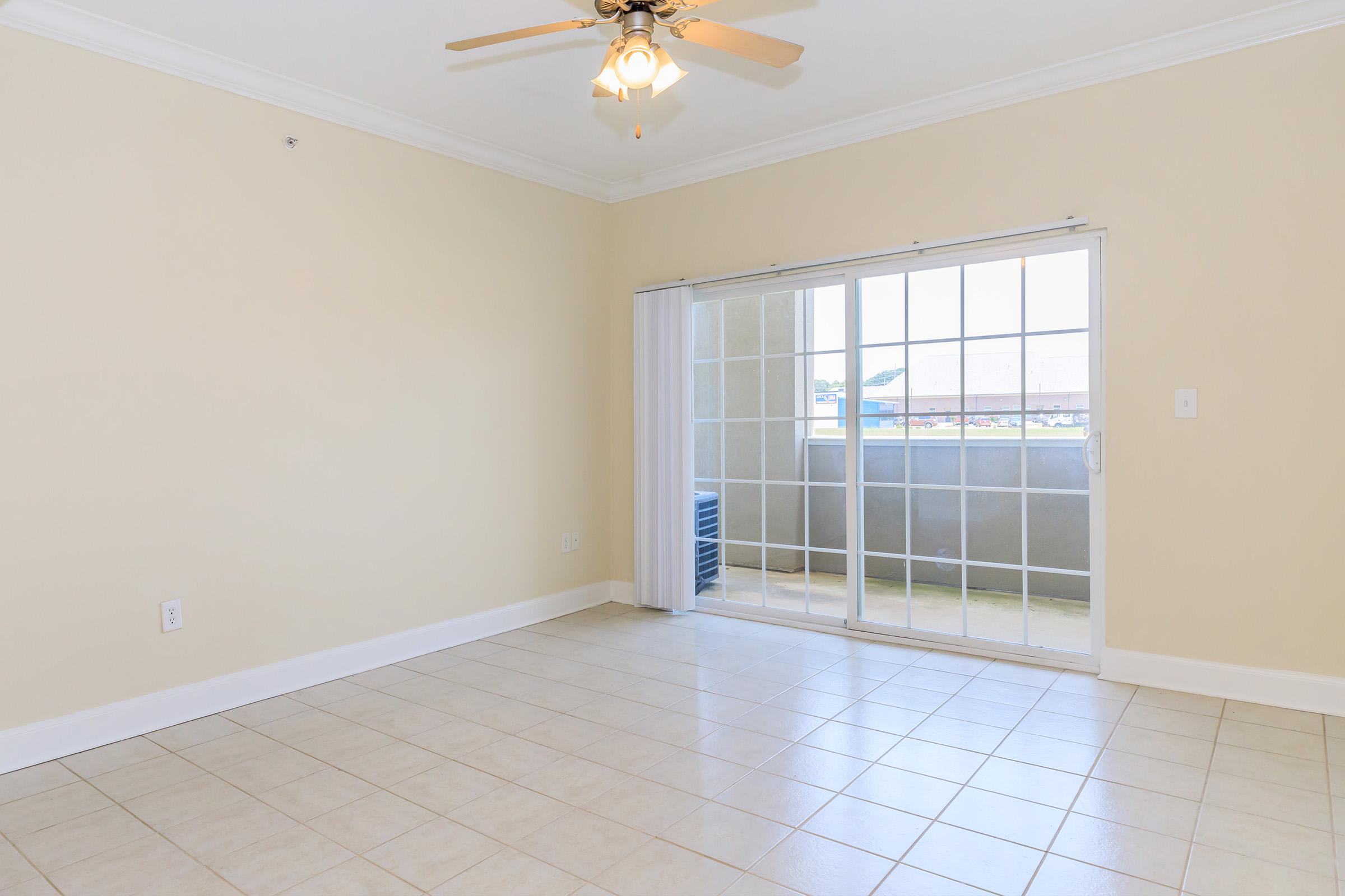 Bright and spacious interior of an empty room with beige walls and tiled floor. A ceiling fan is mounted above, and there is a large sliding glass door that leads to a balcony with a view. Natural light fills the space, creating an inviting atmosphere. No furniture or decor is present.