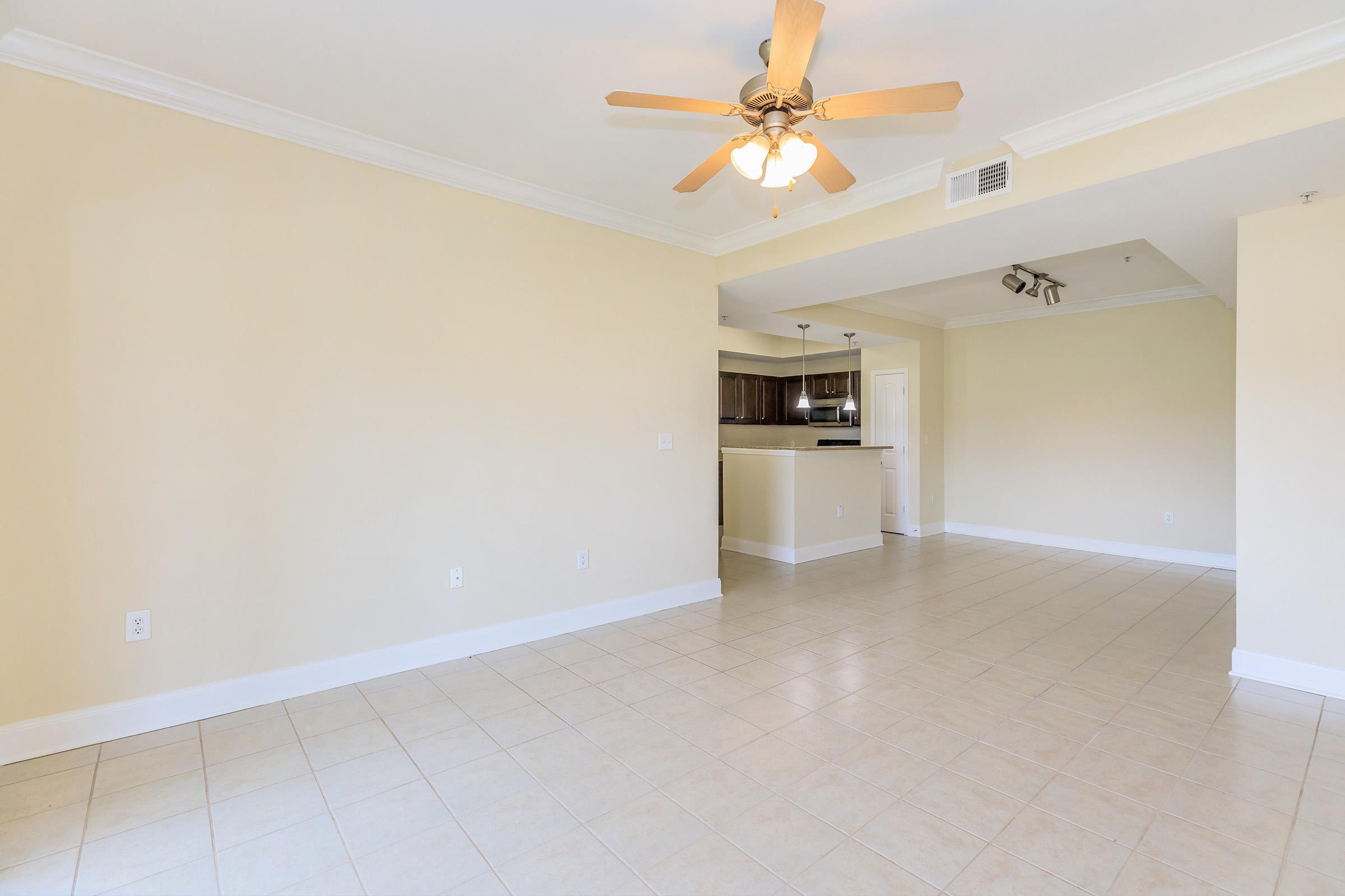 A spacious and well-lit room featuring light-colored walls, a ceiling fan, and tiled flooring. In the background, there is a kitchen area with dark cabinetry. The layout offers a sense of openness, ideal for living and dining purposes. Natural light brightens the space, creating a welcoming atmosphere.