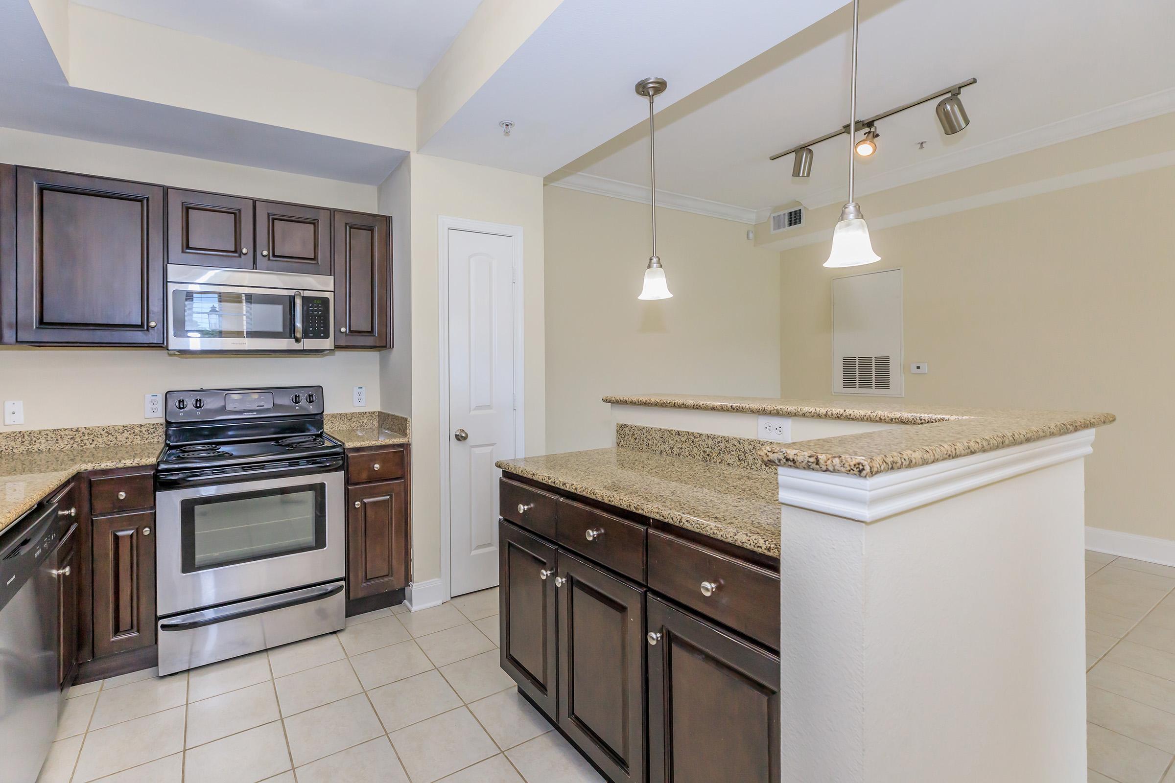 Modern kitchen featuring dark wooden cabinets, stainless steel appliances including a microwave and stove, granite countertops, and pendant lighting. The layout includes a breakfast bar and tile flooring, with a light-colored wall giving a spacious feel. Ideal for a contemporary living space.