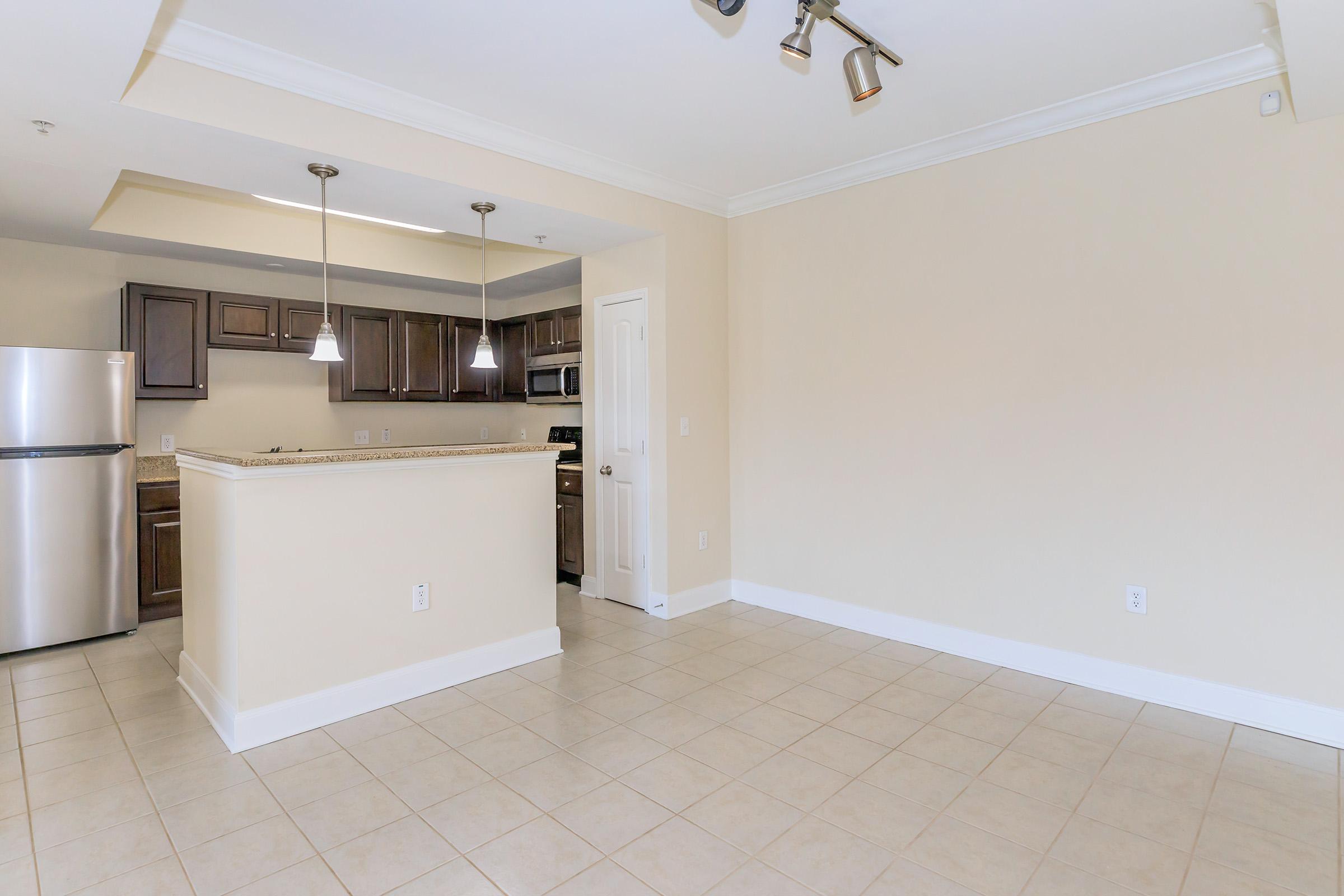 A modern kitchen with dark wooden cabinets and stainless steel appliances, featuring an island with pendant lighting. The room has beige walls, tiled flooring, and an open layout that leads into a spacious area.