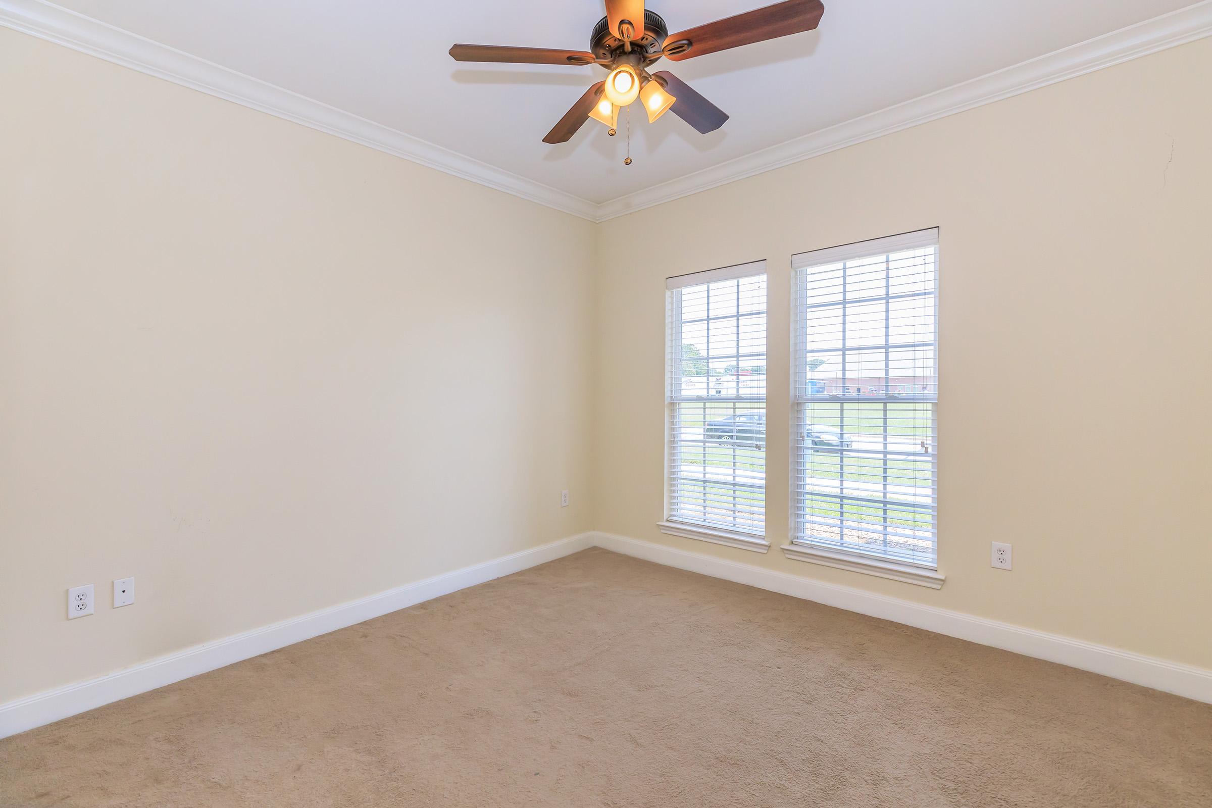 A vacant room with beige walls and carpet. It features a ceiling fan with light fixtures and two windows with white blinds allowing natural light to enter. The space appears clean and well-maintained, ready for furnishing.