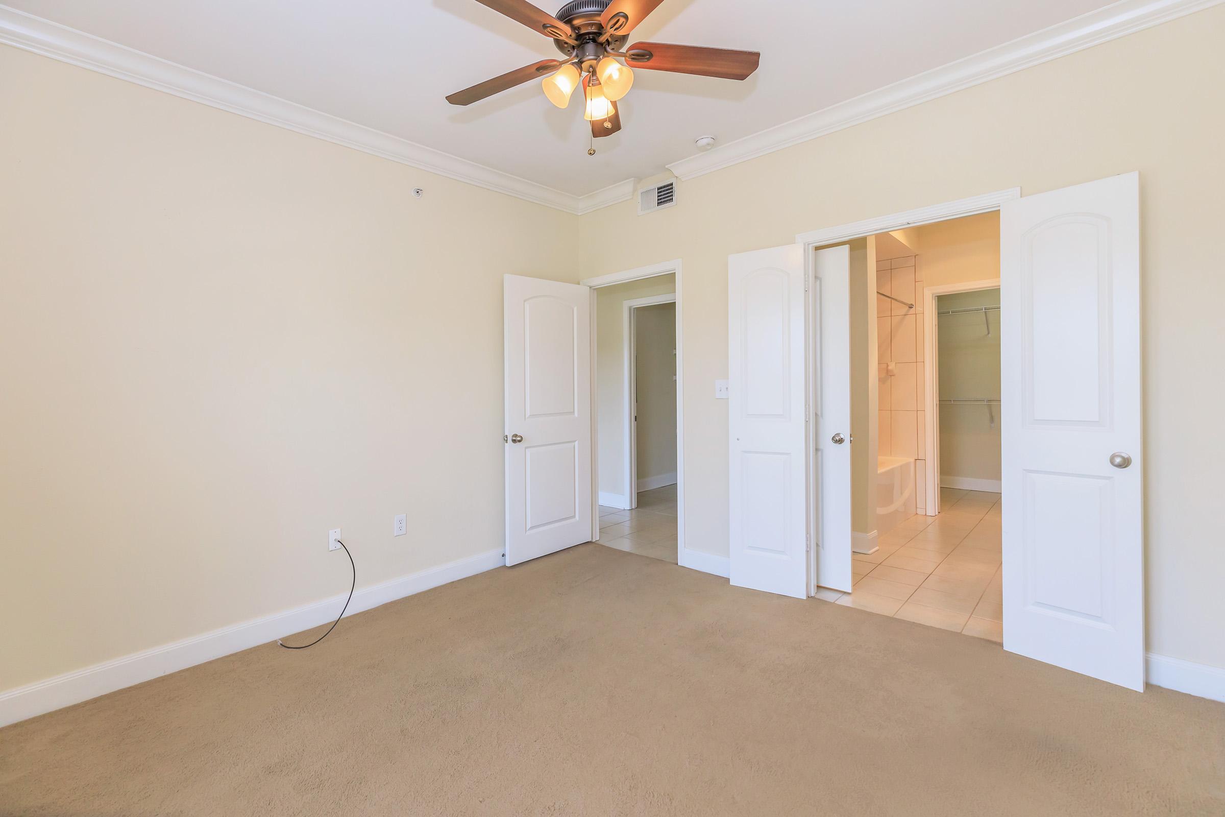 A light-colored room with beige carpet and cream walls. A ceiling fan with wooden blades is visible. Two doors lead to other areas, one possibly to a bathroom, and the room is well-lit by natural light.
