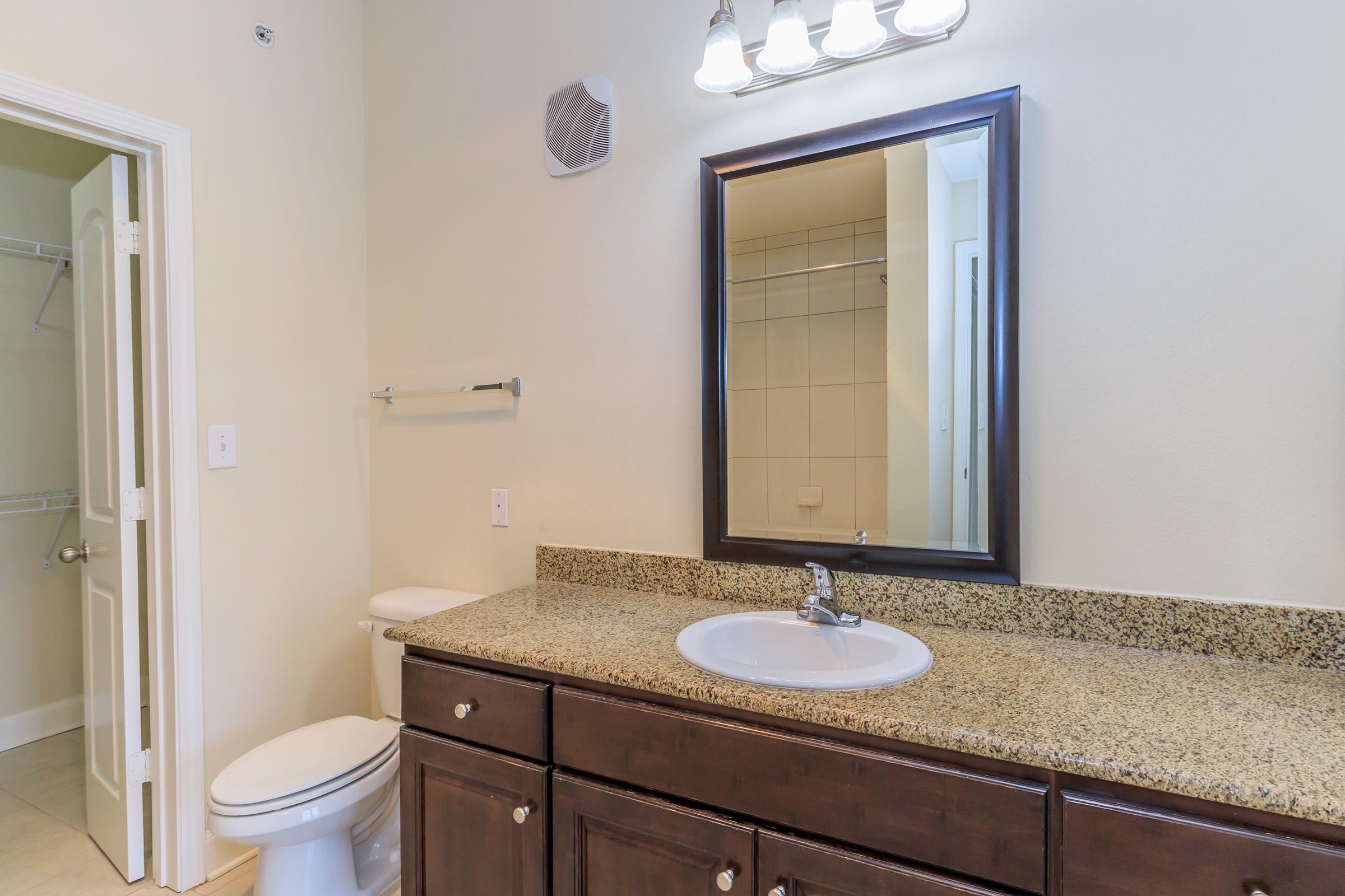 A modern bathroom featuring a granite countertop with a sink, a large mirror above, and a light fixture. The space includes a white toilet and a towel rack, with beige tiled flooring and light-colored walls. A door leads to another room, visible in the background.