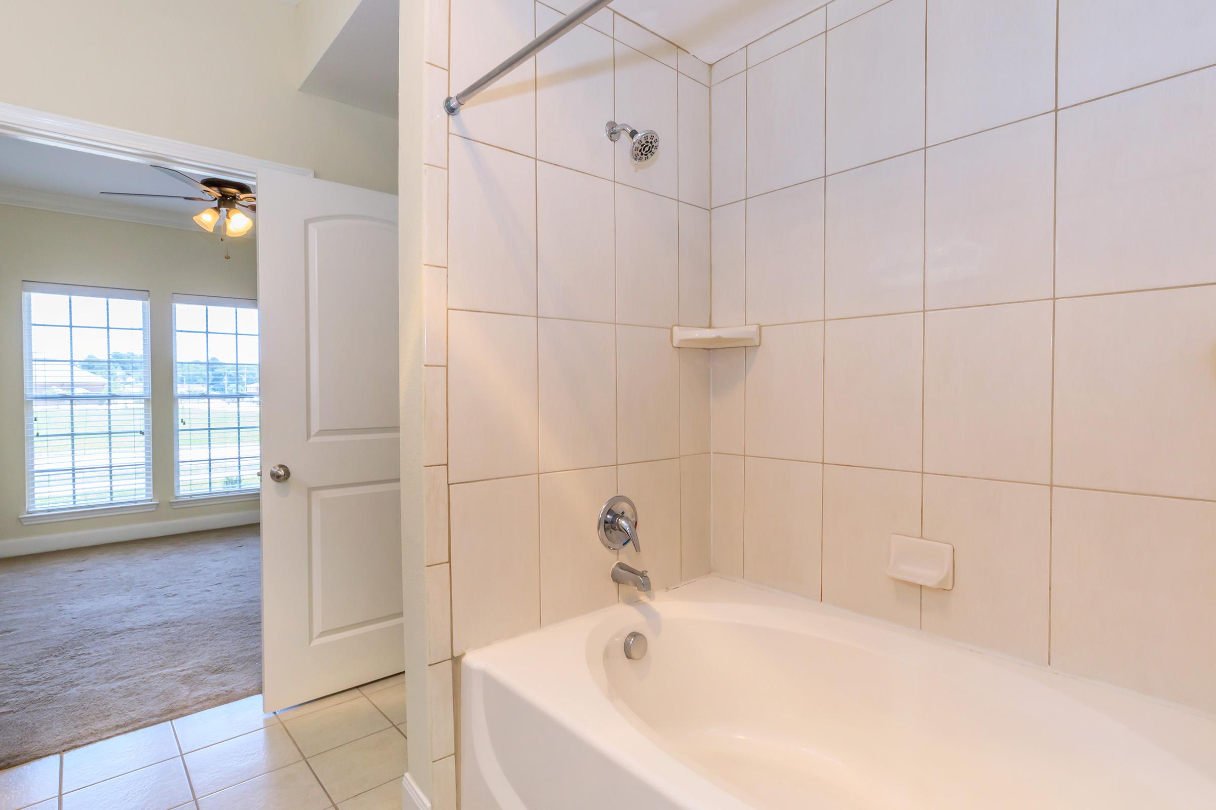A clean and modern bathroom featuring a bathtub with a showerhead, tiled walls, and a shower rod. In the background, a door leads to a brightly lit room with large windows and a carpeted floor.