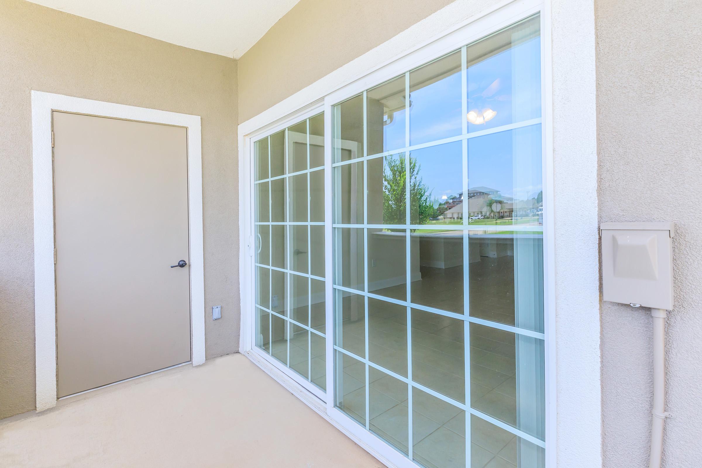 A view of a patio area featuring a large sliding glass door with a grid pattern, leading to an interior space. The door is framed by a beige exterior wall and a simple brown door is visible nearby. Outside, there's a glimpse of a landscaped area beyond the glass door.