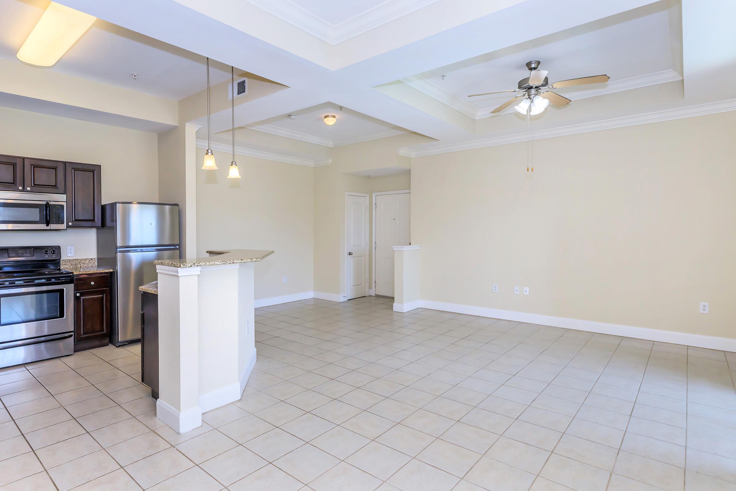 A spacious, empty living area featuring light-colored walls and a ceiling fan. The kitchen area, with dark wooden cabinets, stainless steel appliances, and a granite countertop, is partially visible. Large tiles cover the floor, creating an open and airy feel with ample natural light.