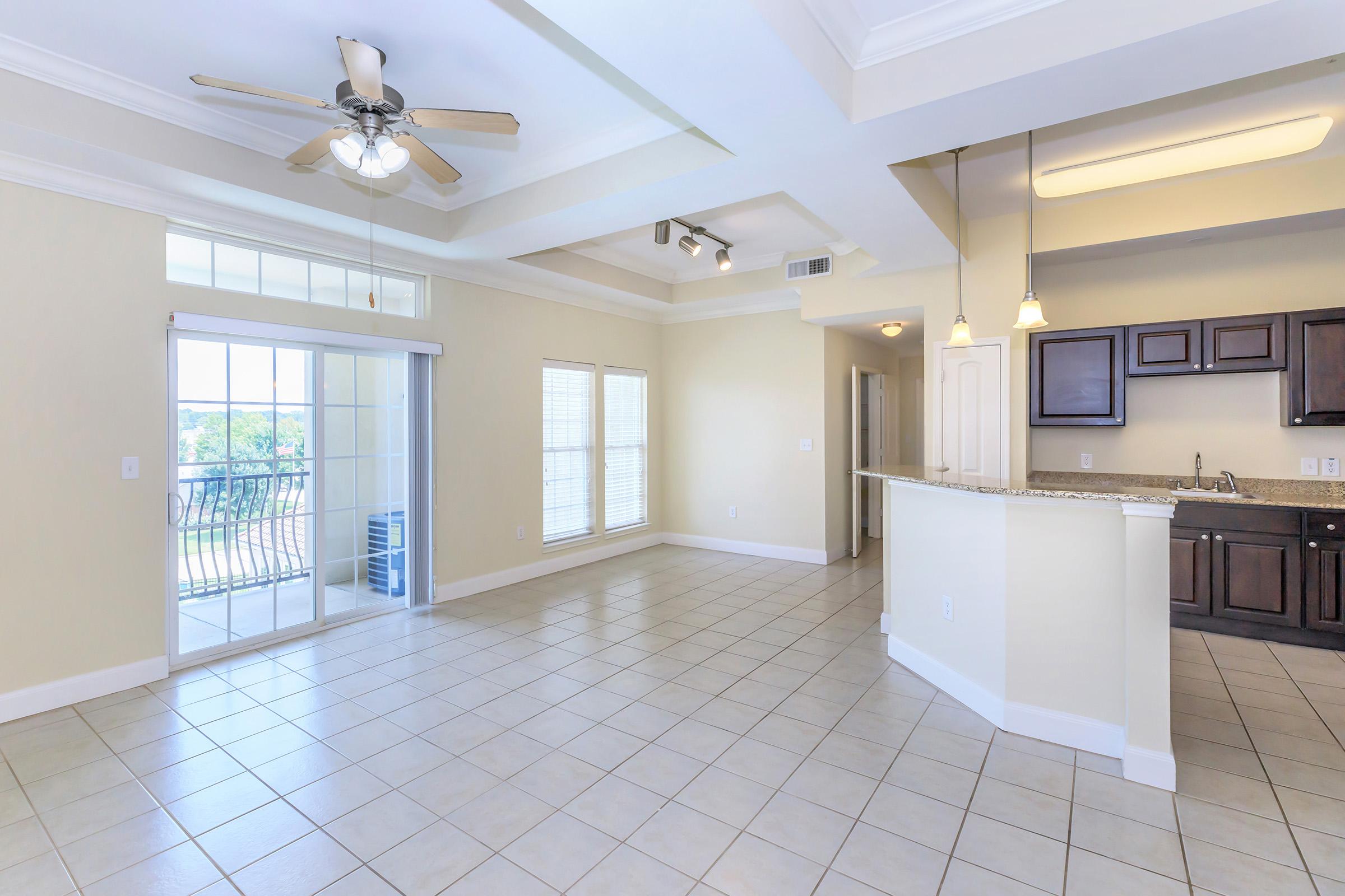 Spacious living area in a modern apartment featuring beige walls, tiled floor, and a ceiling fan. There are large windows with blinds allowing natural light, and an open kitchen with dark wood cabinets and a granite countertop. A balcony door leads to an outdoor space.