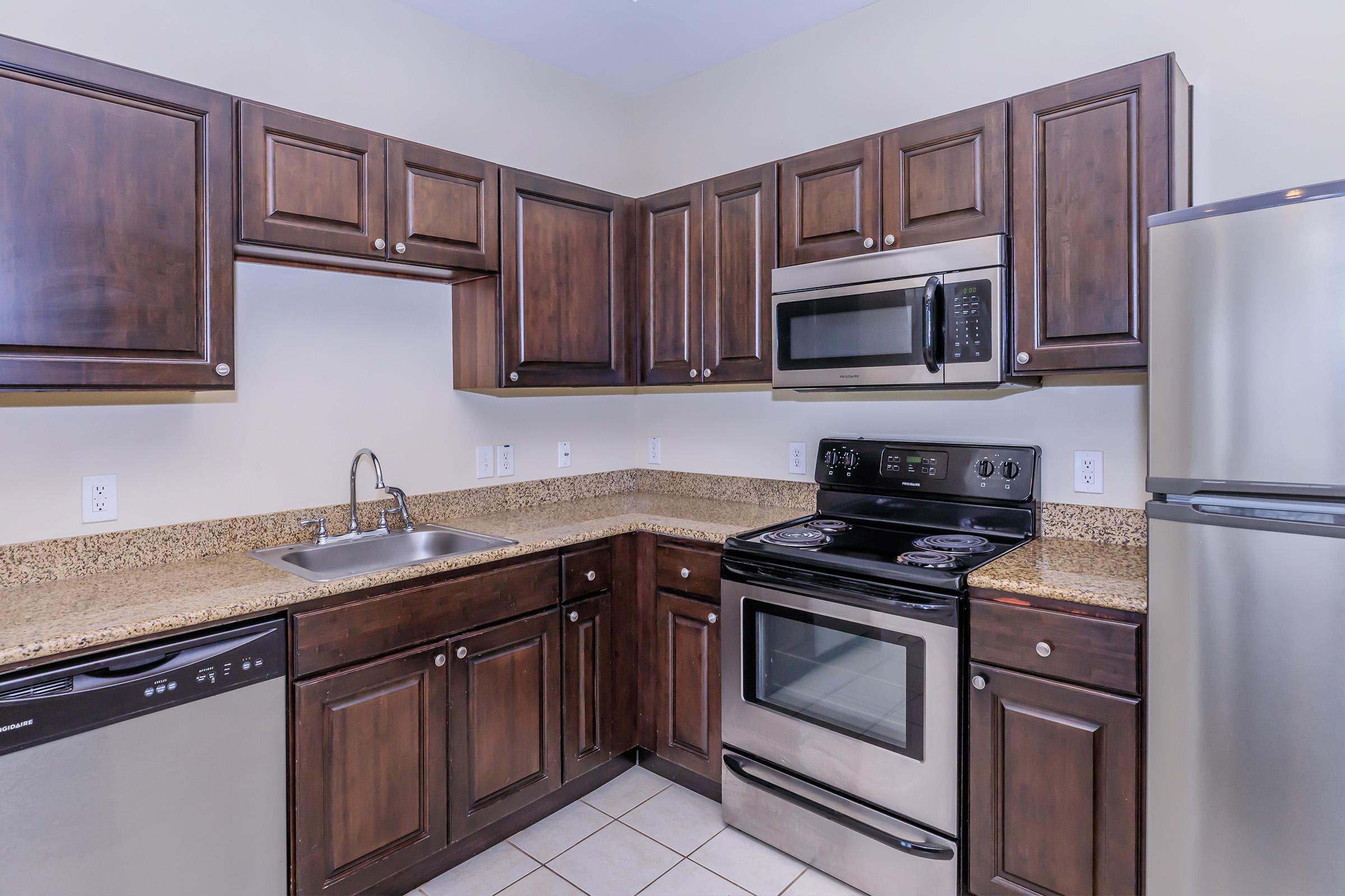 Modern kitchen featuring dark wooden cabinets, granite countertops, stainless steel appliances including an oven, microwave, and refrigerator, and a single-basin sink. The space has tiled flooring and a neutral wall color, creating a sleek and contemporary look.