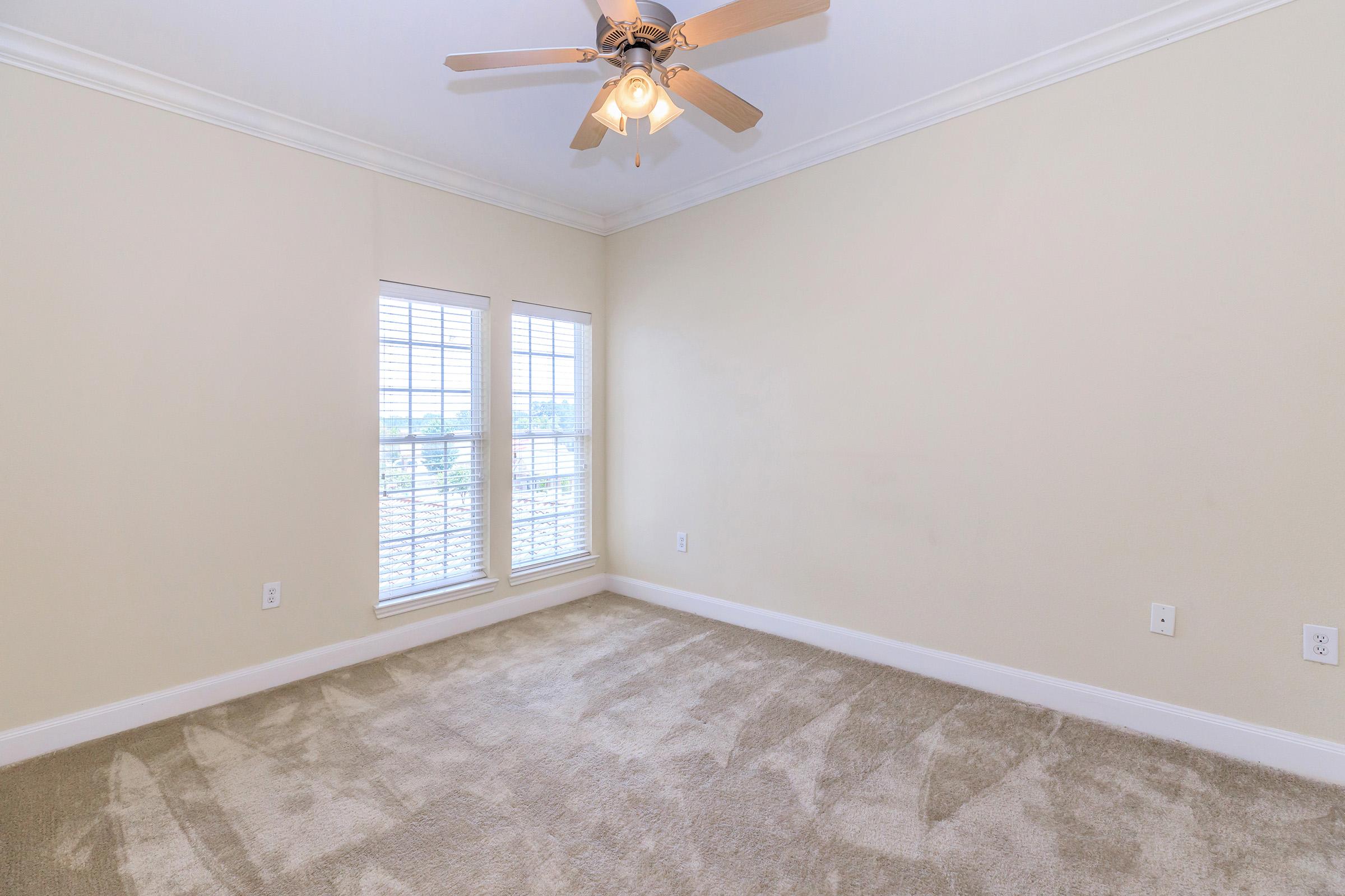 Empty room featuring light-colored walls, a ceiling fan with light fixture, and two windows with shutters. The floor is covered in beige carpet, and there is a view of the outdoors visible through the windows.