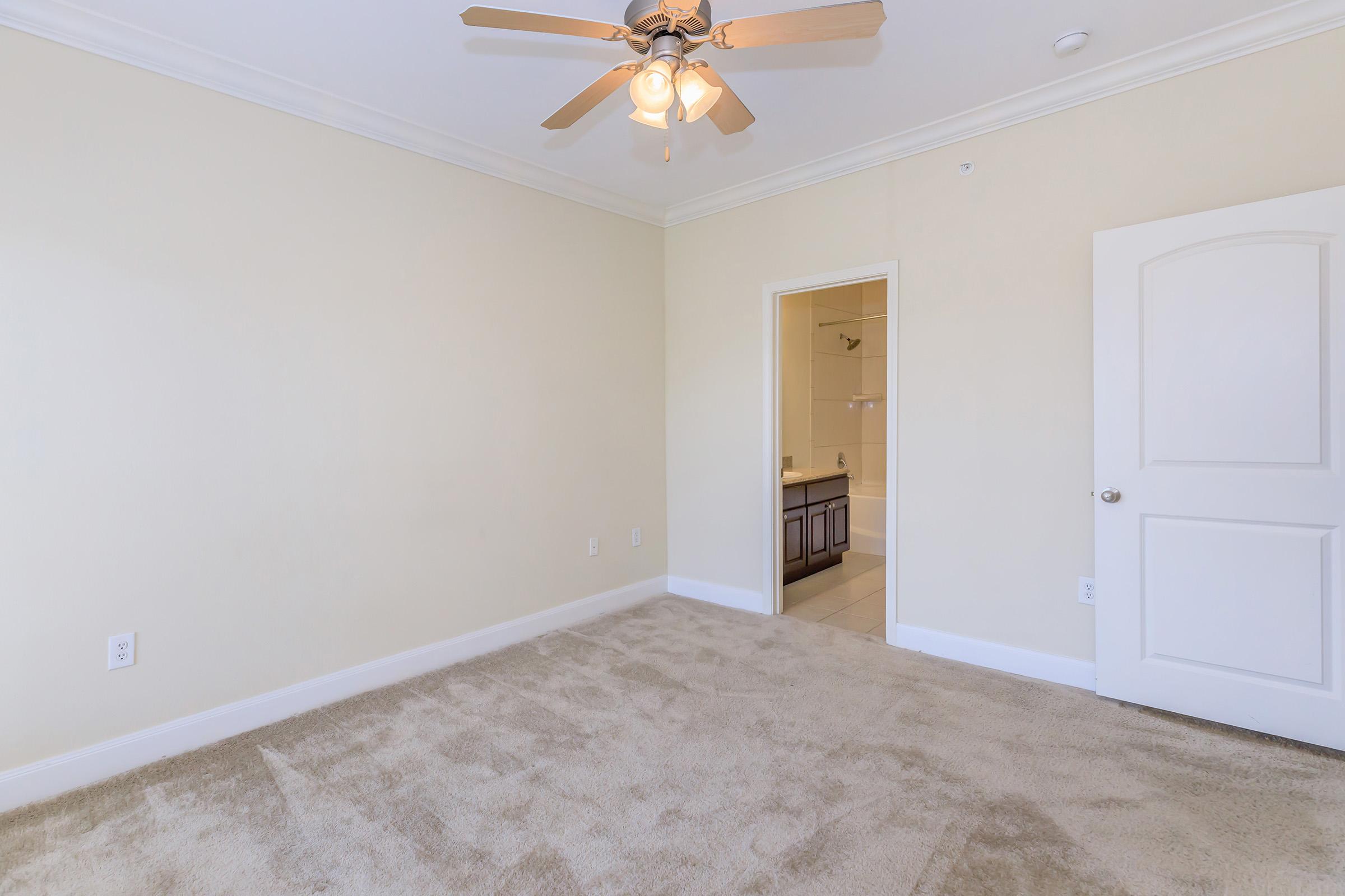 A spacious room with beige walls and a light-colored carpet. There is a ceiling fan with wooden blades, and a doorway leading to an adjacent bathroom. Natural light fills the space, creating a warm and inviting atmosphere.