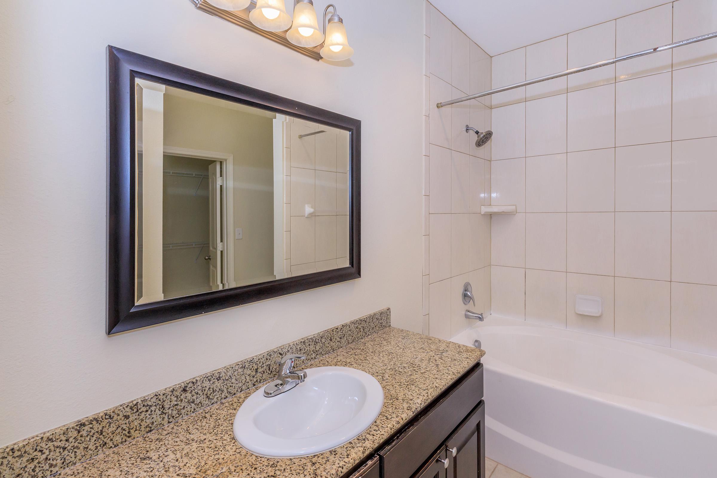 Modern bathroom featuring a granite countertop with a sink, a large mirror above, and a bathtub with a shower. The walls are tiled in neutral colors, and there is a light fixture with multiple bulbs above the mirror. The design is clean and contemporary.