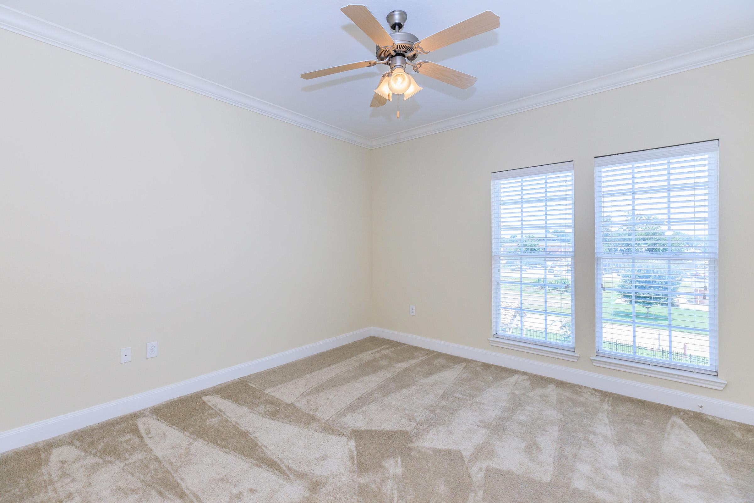 Empty room with light beige walls and carpet. A ceiling fan with wooden blades is mounted in the center. Two windows with white blinds allow natural light to enter. The space appears clean and spacious, ideal for potential furnishing and decoration.