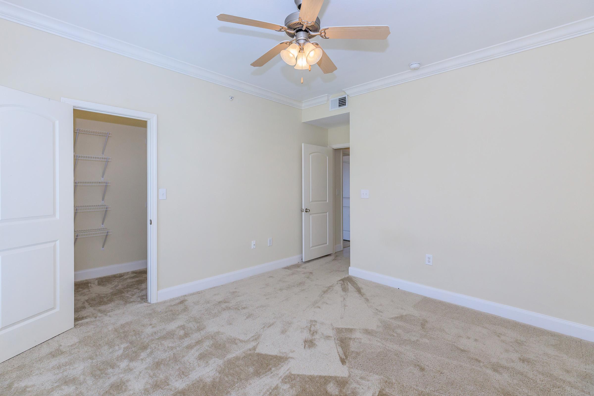 A spacious room with light beige walls and carpet, featuring a ceiling fan with wooden blades. There is an open door leading to a closet on the left and another door to a bathroom or hallway on the right. The room is well-lit with natural light coming from an unseen window.