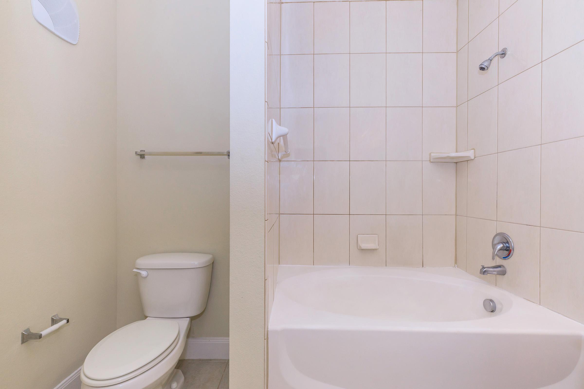A clean bathroom featuring a white toilet, a bathtub with a shower, and tiled walls. The space is well-lit with neutral colors, and there are grab bars installed for safety.