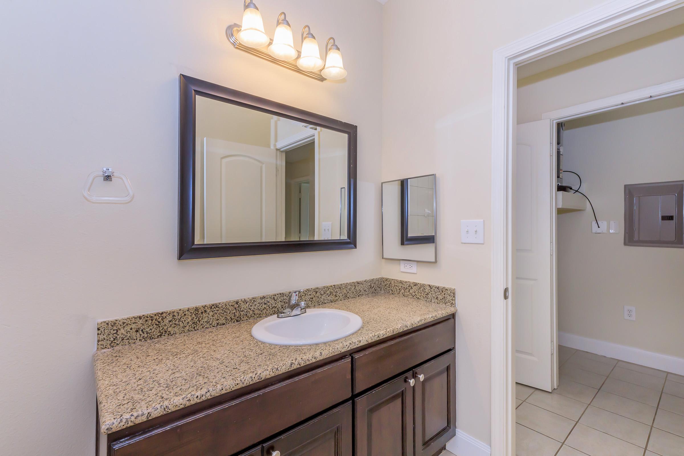 A well-lit bathroom with a large mirror above a granite countertop. Below the counter are dark wood cabinets. There is a towel holder mounted on the wall and a small mirror beside the main mirror. A door leads to a utility room, and the flooring is tiled.