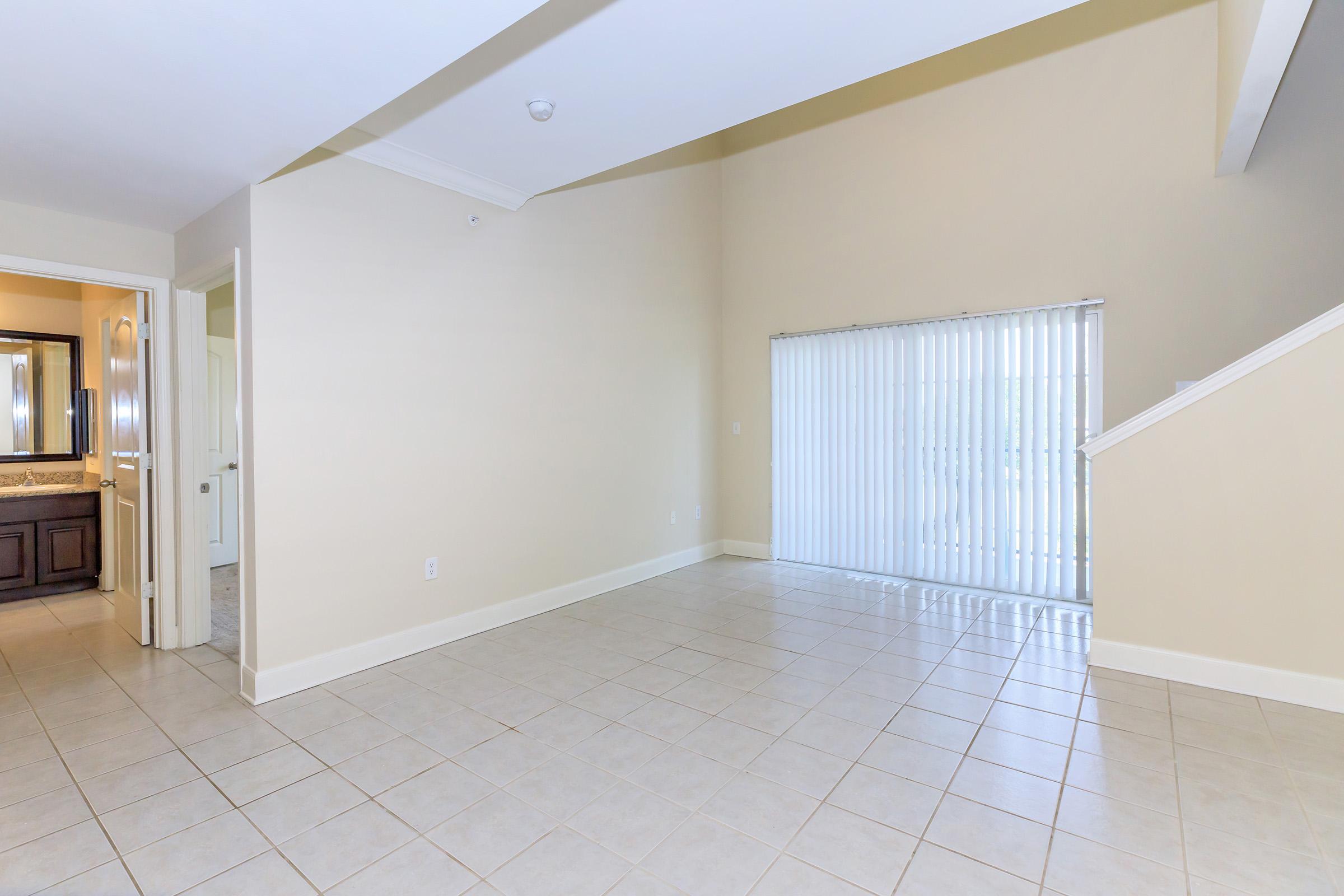 A spacious, empty interior room with beige walls and tiled flooring. On the left, a doorway leads to a bathroom with dark wood cabinetry. A large window with vertical blinds on the right allows natural light to enter, and a staircase is visible in the background, leading to the upper level.