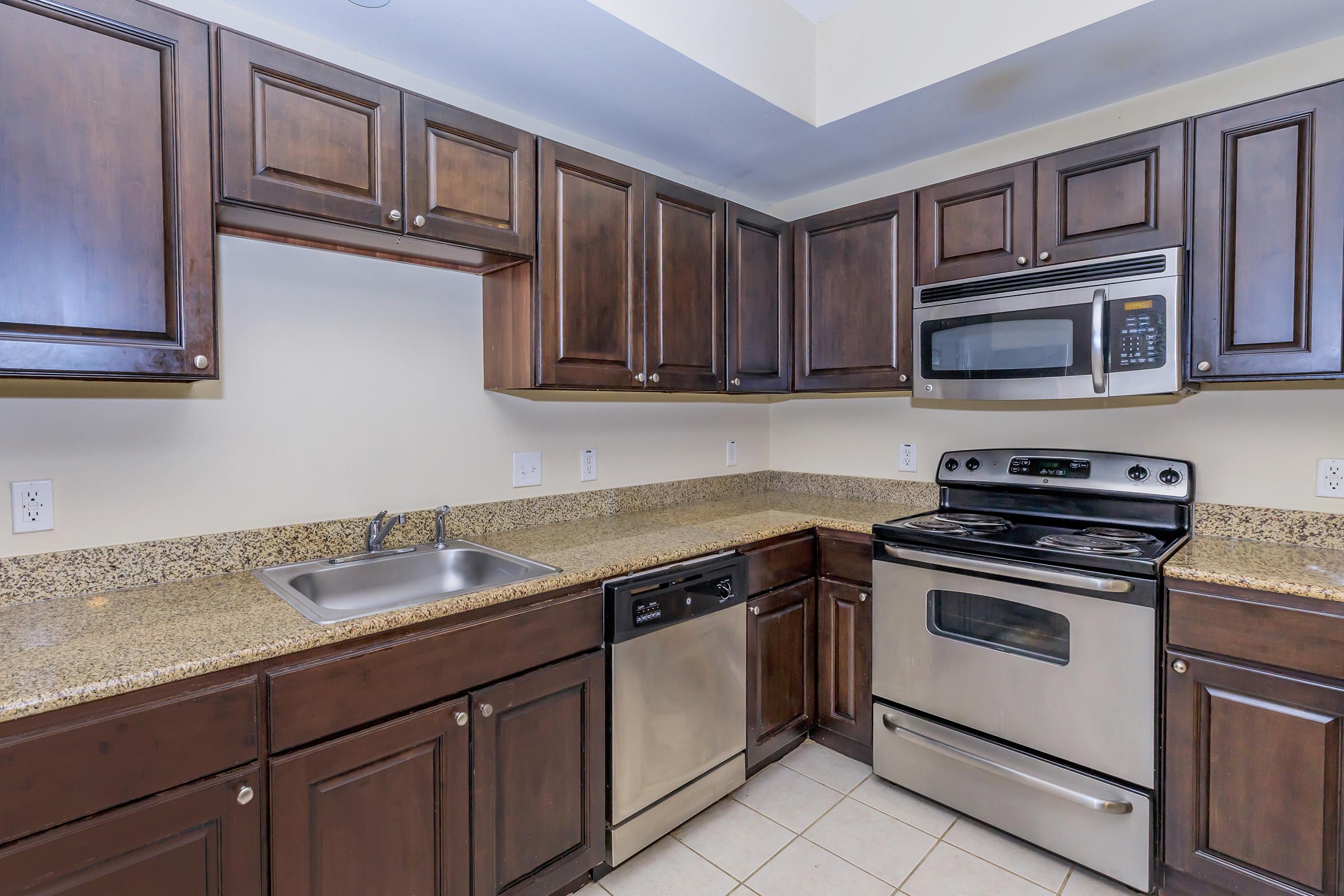 Modern kitchen featuring dark wood cabinets, granite countertops, stainless steel appliances including an oven, microwave, dishwasher, and sink. The design is clean and functional, with tile flooring and a neutral wall color, creating an inviting cooking space.