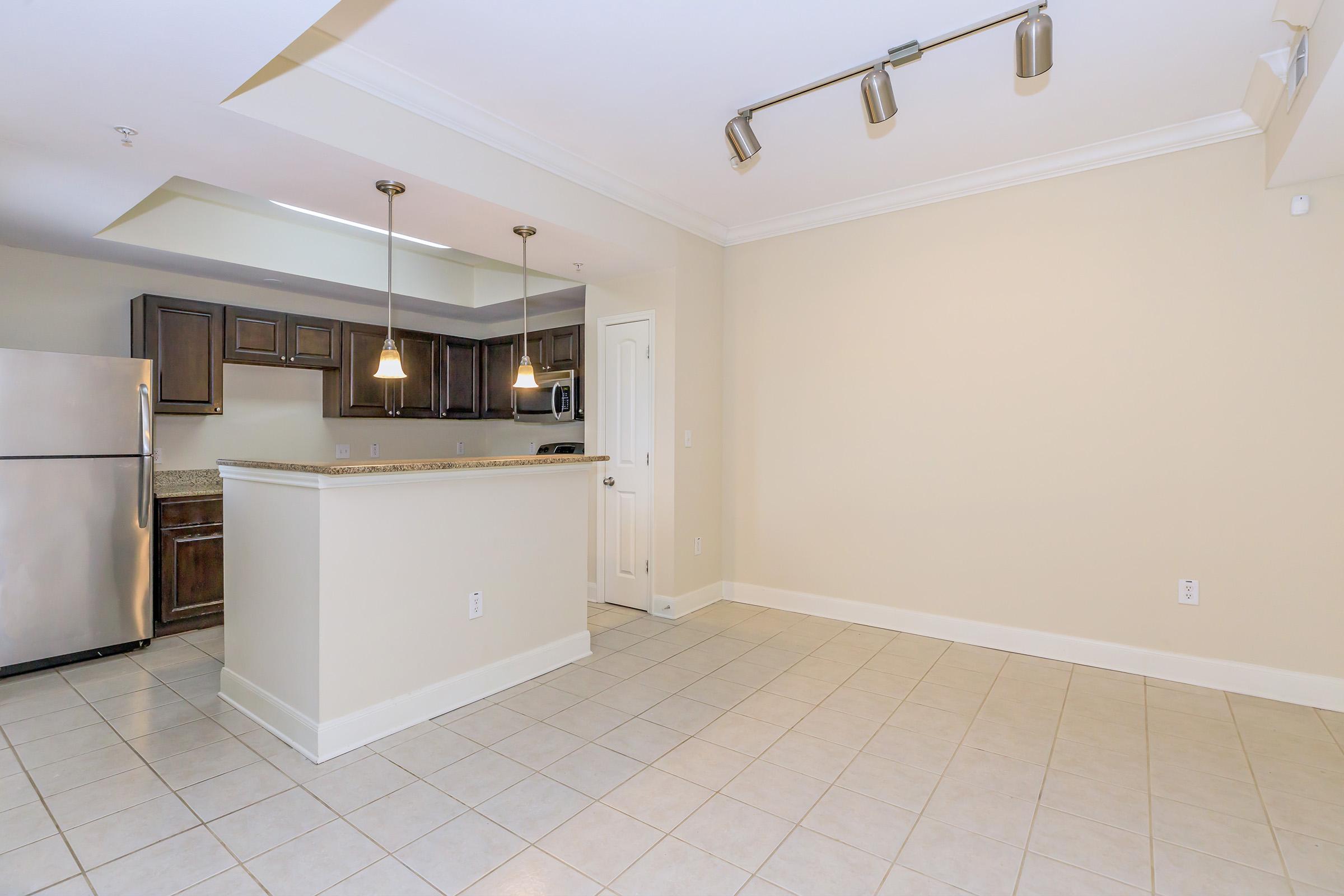 Modern kitchen and living area showing a spacious layout. The kitchen features dark wood cabinets, stainless steel appliances, and pendant lighting over the island. The floor is tiled, and the walls are painted a light beige color, creating a warm and inviting atmosphere.