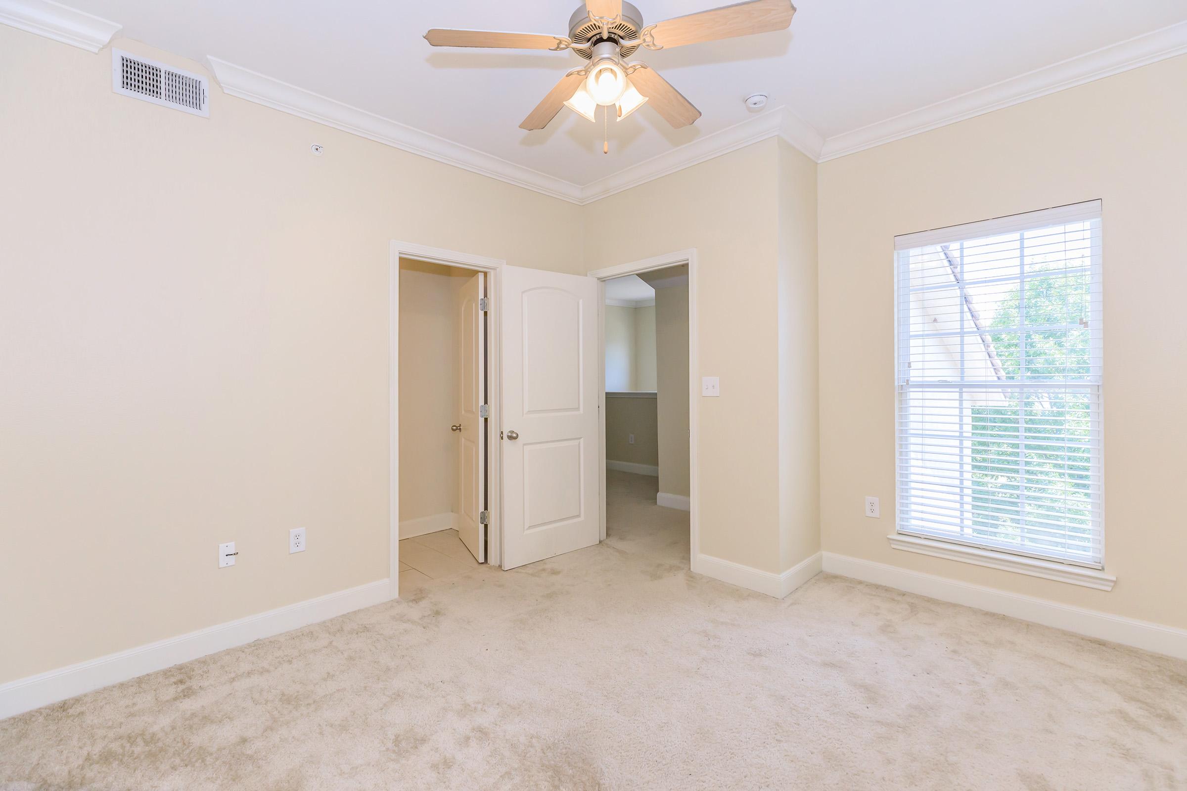 A light and airy bedroom with beige walls and plush carpet. A ceiling fan is mounted in the center, and there is a large window with blinds letting in natural light. Two doors lead to adjacent rooms, one slightly ajar, creating an inviting and cozy atmosphere.
